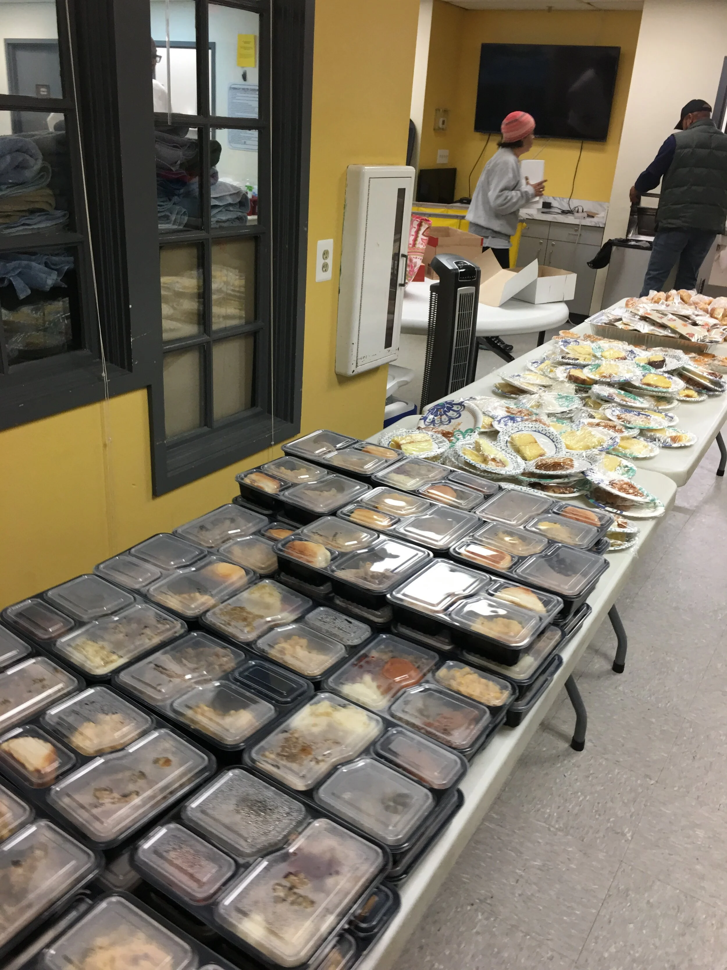 Table filled with pre-packaged meals and plates of baked goods in a community kitchen or cafeteria setting.