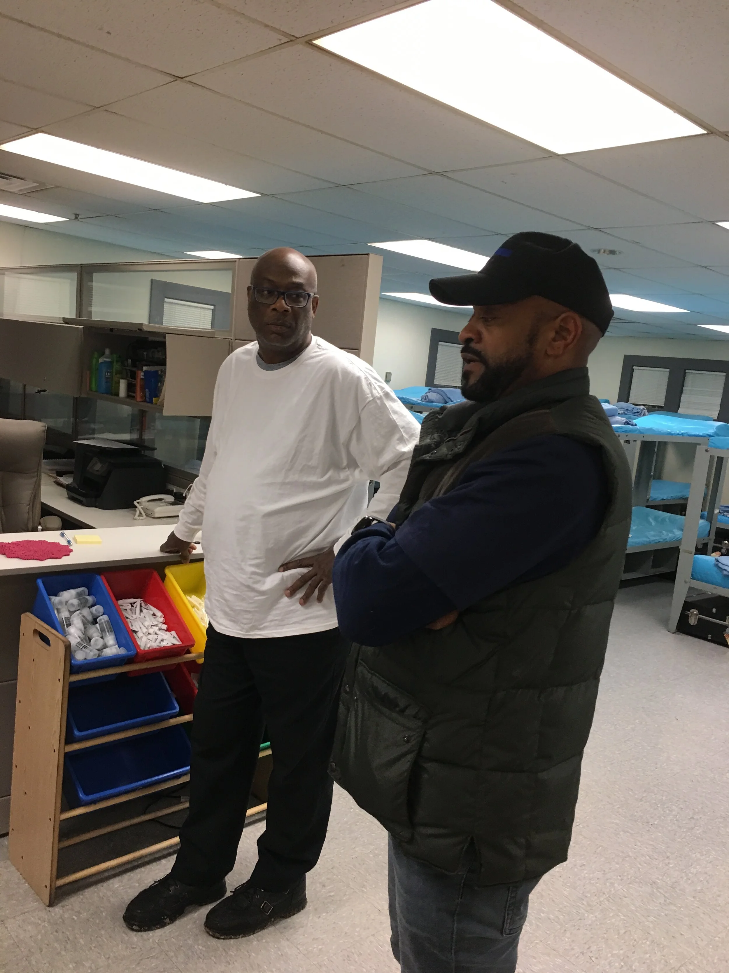 Two men are standing in a room with residential-style bunk beds. One man is wearing a white long-sleeve shirt and glasses, and the other man is wearing a black baseball cap and a black vest. They appear to be engaged in conversation.