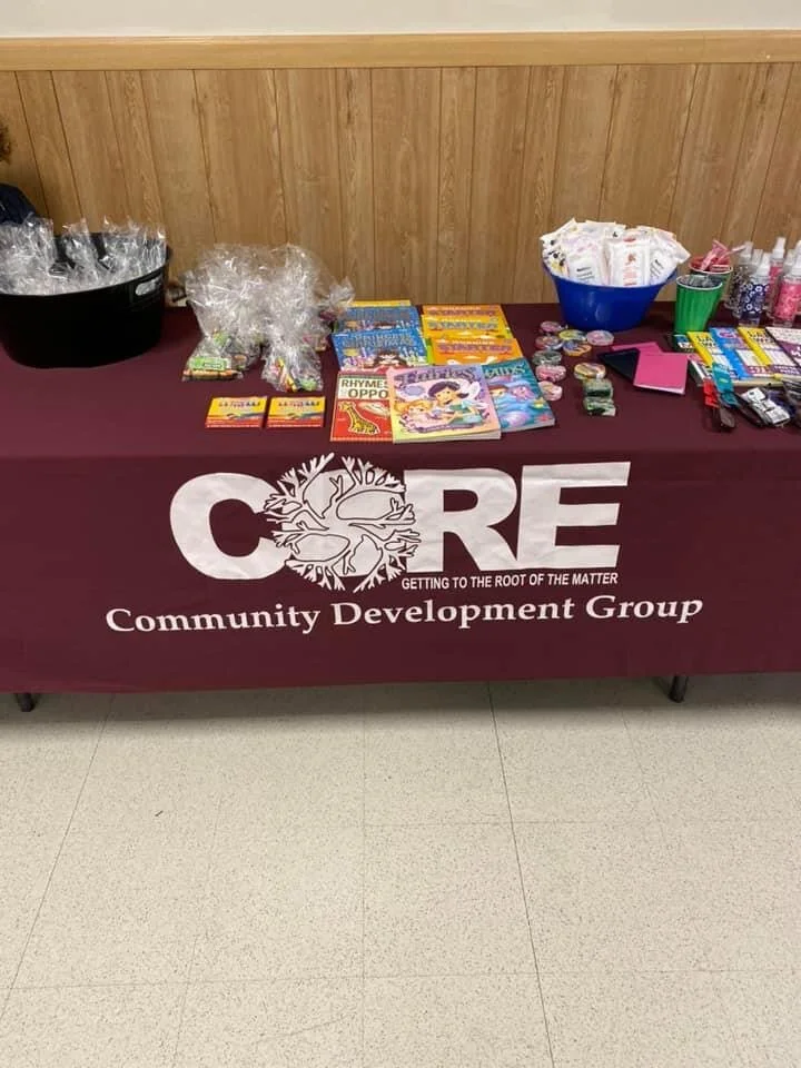 A table with various school supplies, snacks, and small toys or candies. The table has a maroon cloth with the text 'CORE Community Development Group' and a logo featuring a tree, along with the tagline 'Getting to the root of the matter'.