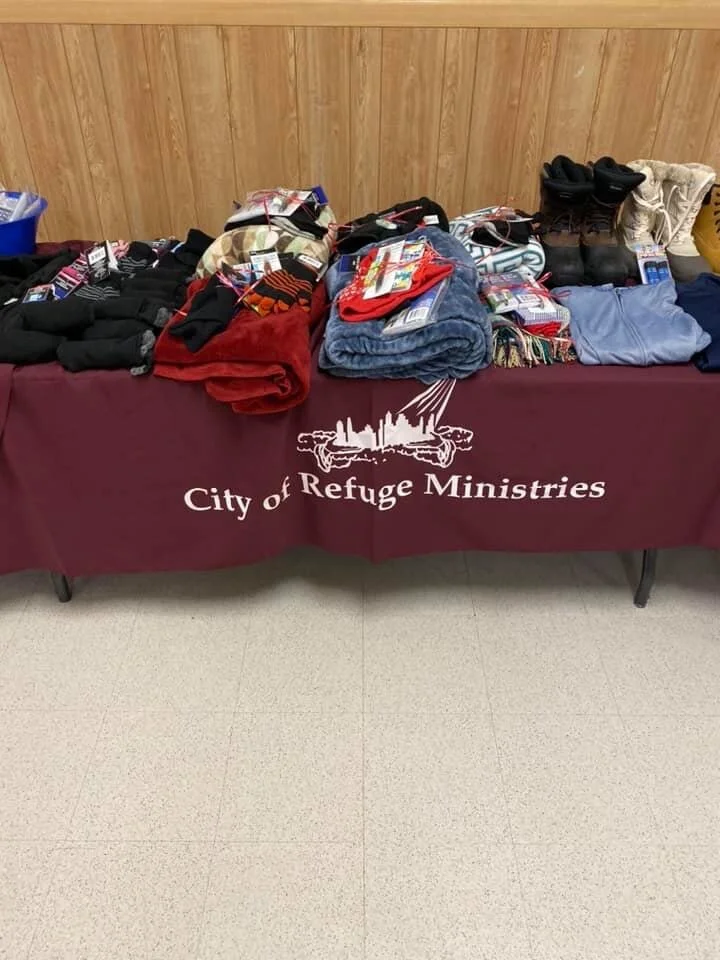 Table with neatly folded clothing, shoes, and accessories, covered by a maroon tablecloth that has the logo and text 'City of Refuge Ministries' printed on it.
