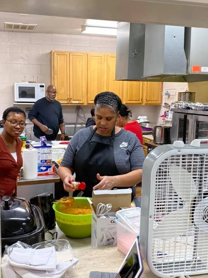 Group of people in a kitchen preparing food, with one woman pouring spices into a large green mixing bowl.