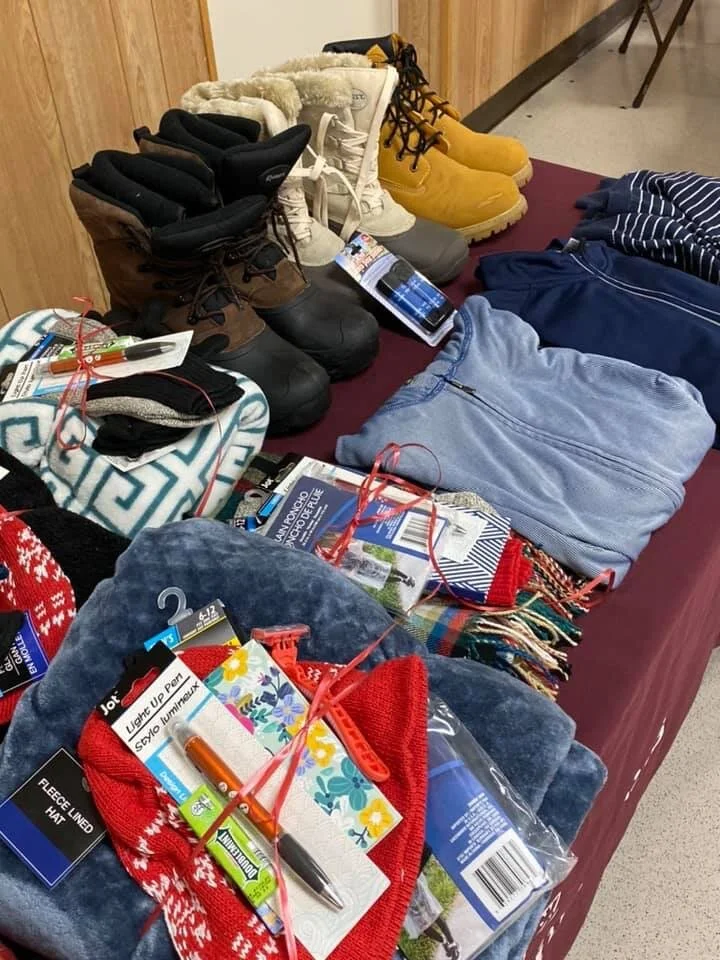 A table displaying new winter boots, folded clothes, scarves, and holiday items, including gift tags and wrapping supplies, at a retail store.