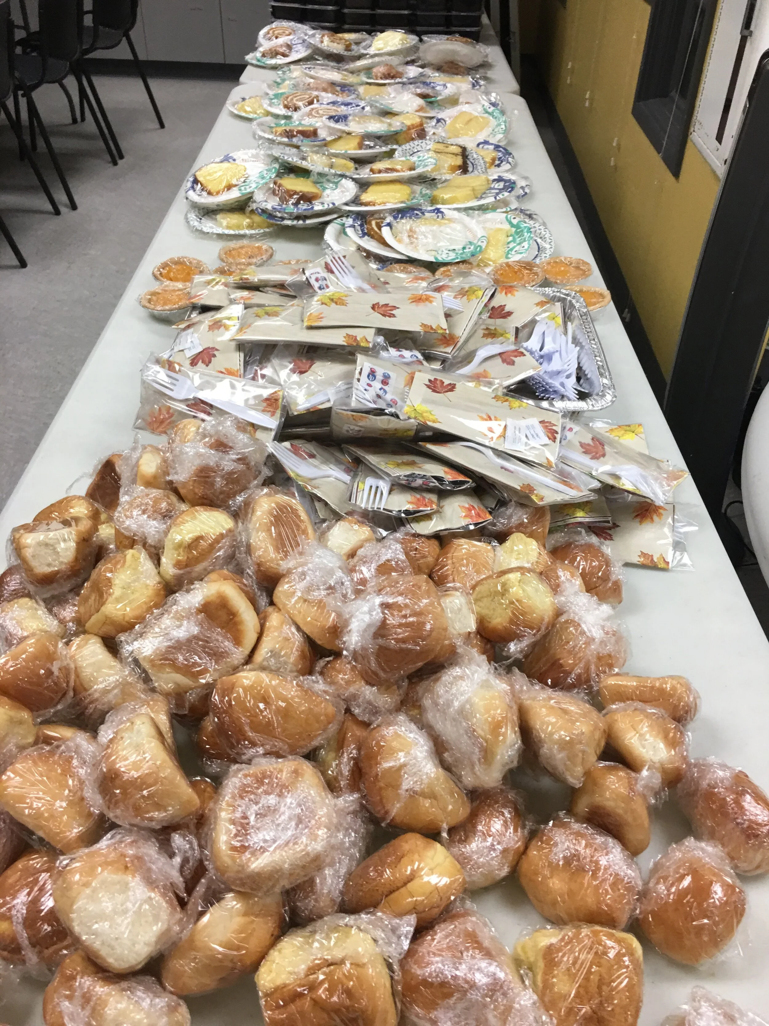 A long table filled with baked goods and snacks, including individual bread rolls wrapped in plastic, packets of chips, small containers of pudding, and plates of assorted pastries, set up in a room with chairs and a yellow wall.