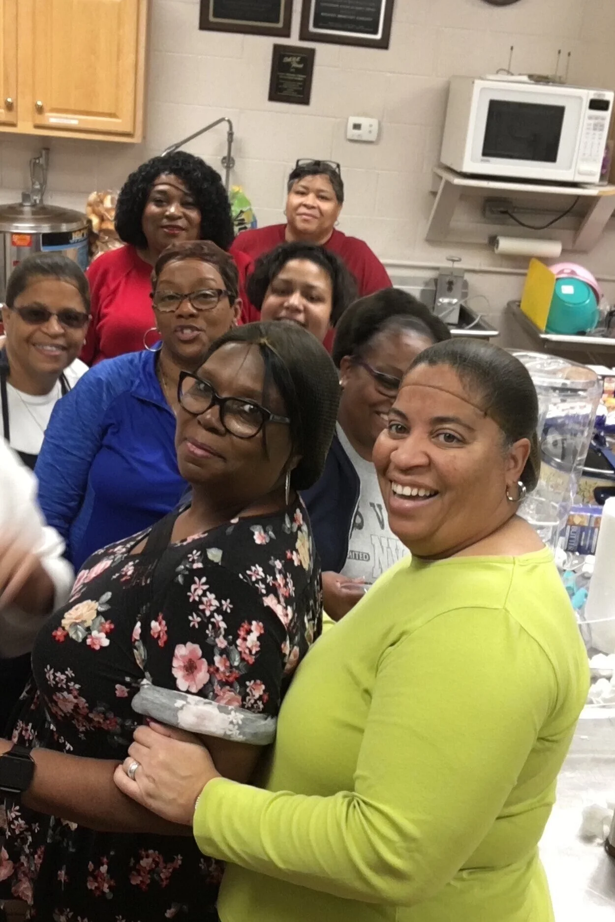 Group of diverse women smiling and posing together in a kitchen or break room.