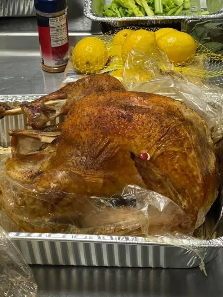 Roast turkey in a metal pan with lemons, greens, and seasoning in the background.