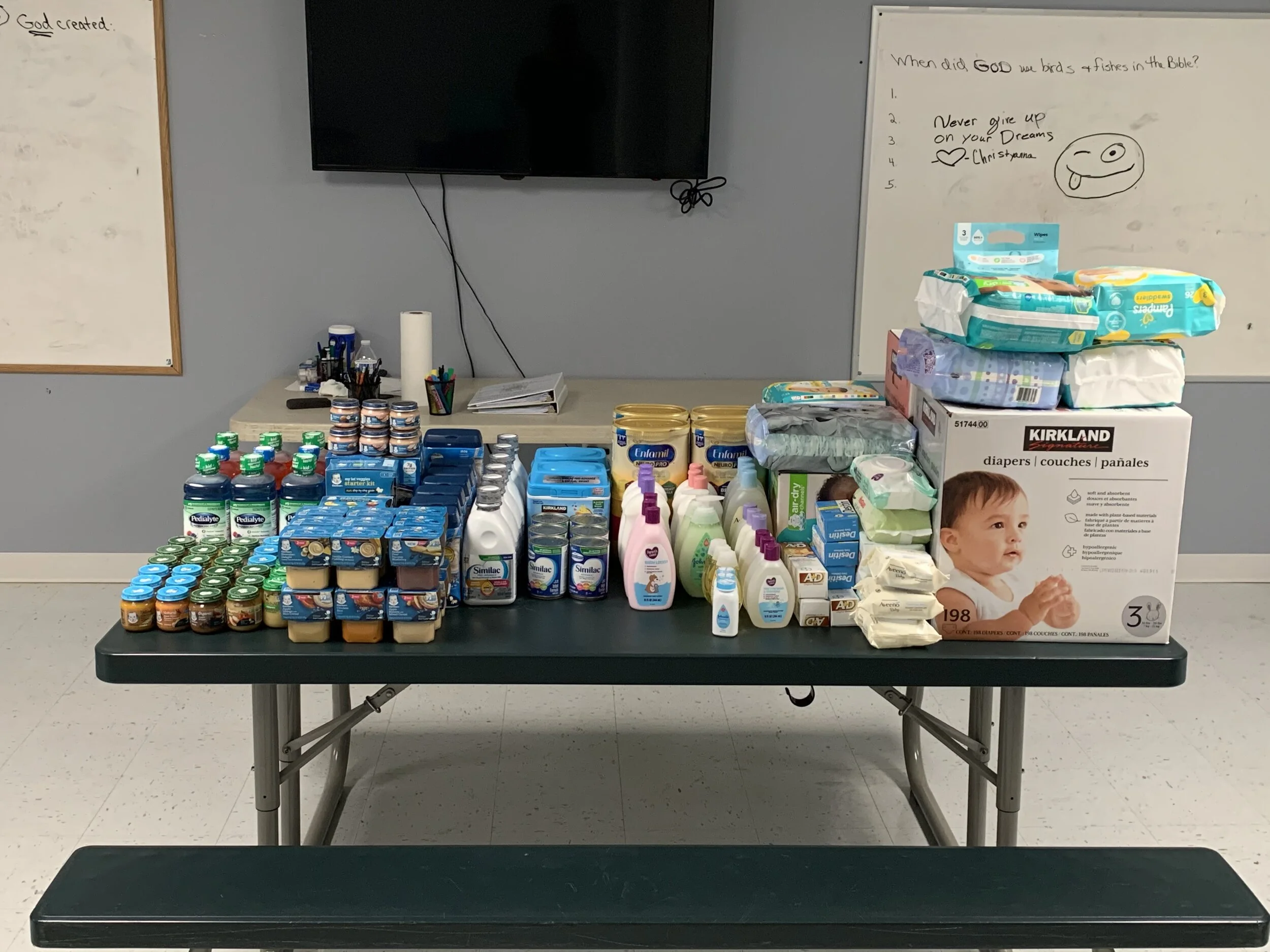 Table filled with various baby care products including bottles, jars, pouches, and boxes, with a large box of diapers on the right side. A whiteboard and a television are visible in the background.