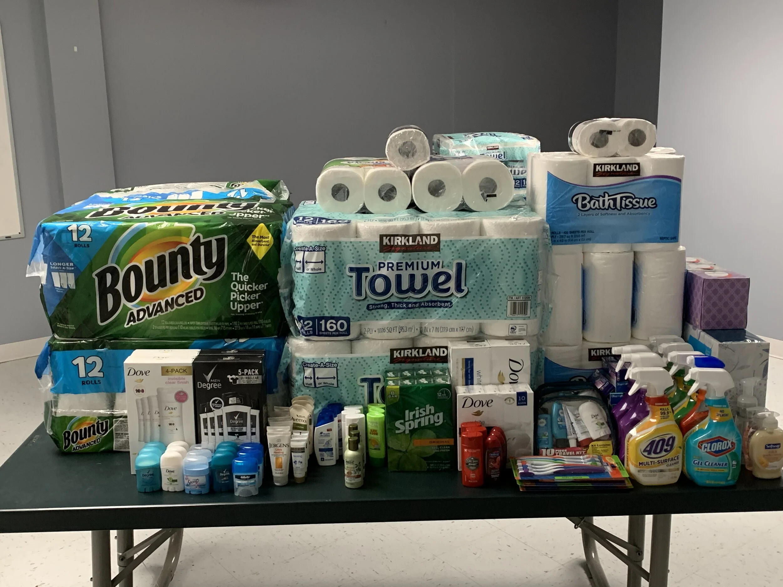 A table filled with household supplies including toilet paper, paper towels, tissue boxes, personal care products, and cleaning sprays.