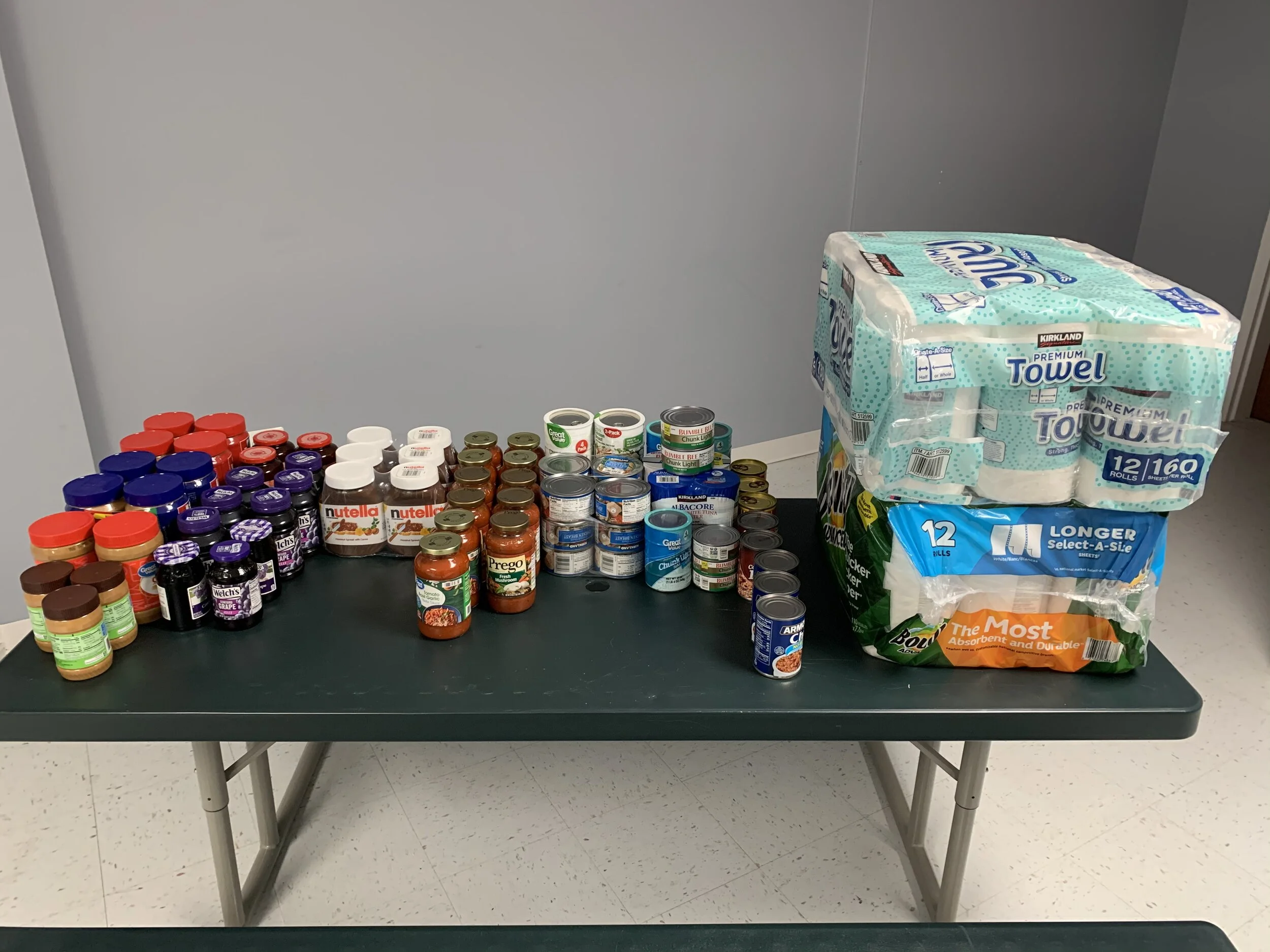 Table with various canned and jarred food items, including Nutella jars, canned vegetables, and soup cans, next to large packs of paper towels and paper napkins.