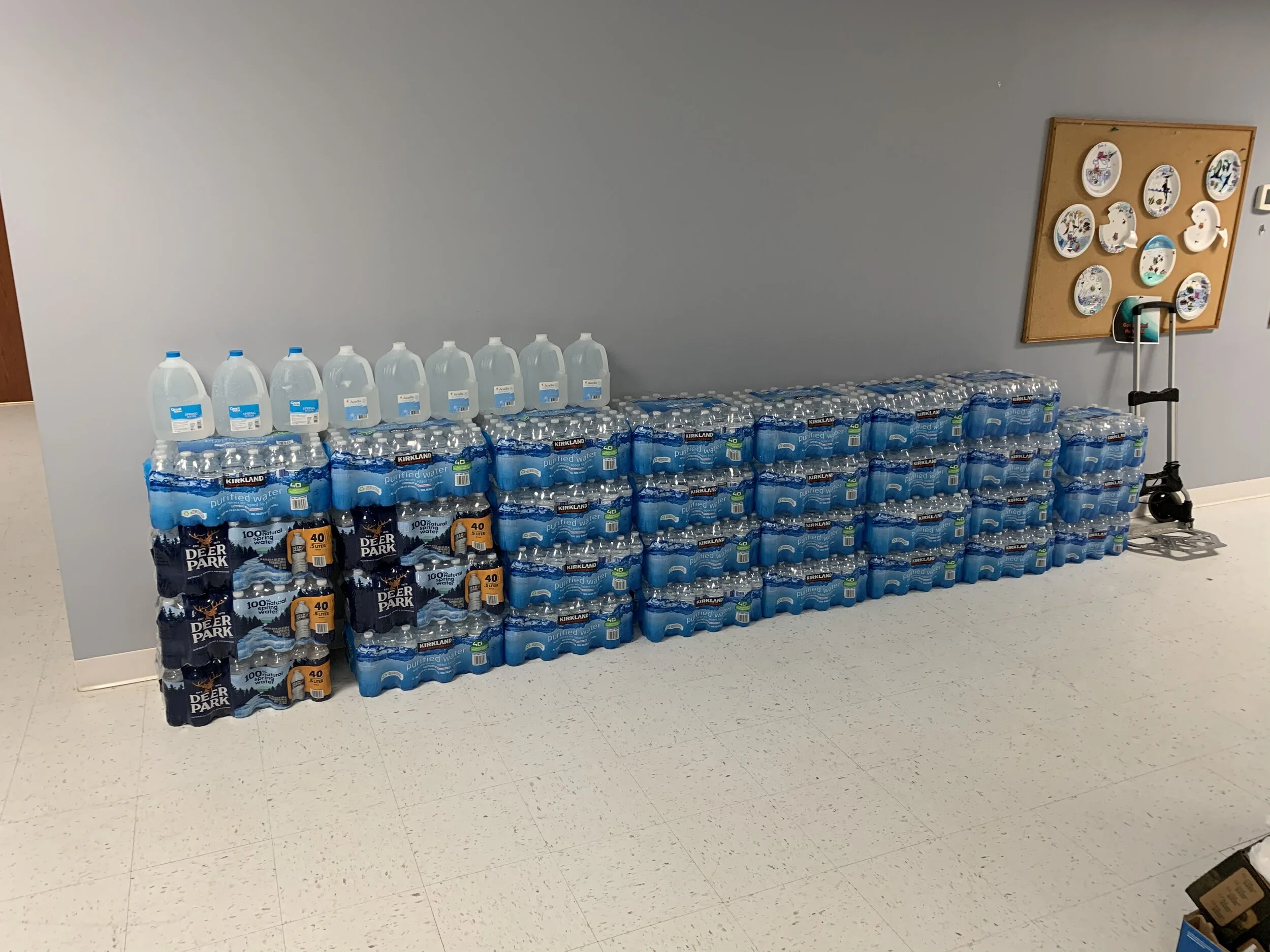 Large quantity of bottled water stacked on a table against a gray wall, with a cane scooter and a bulletin board in the background.
