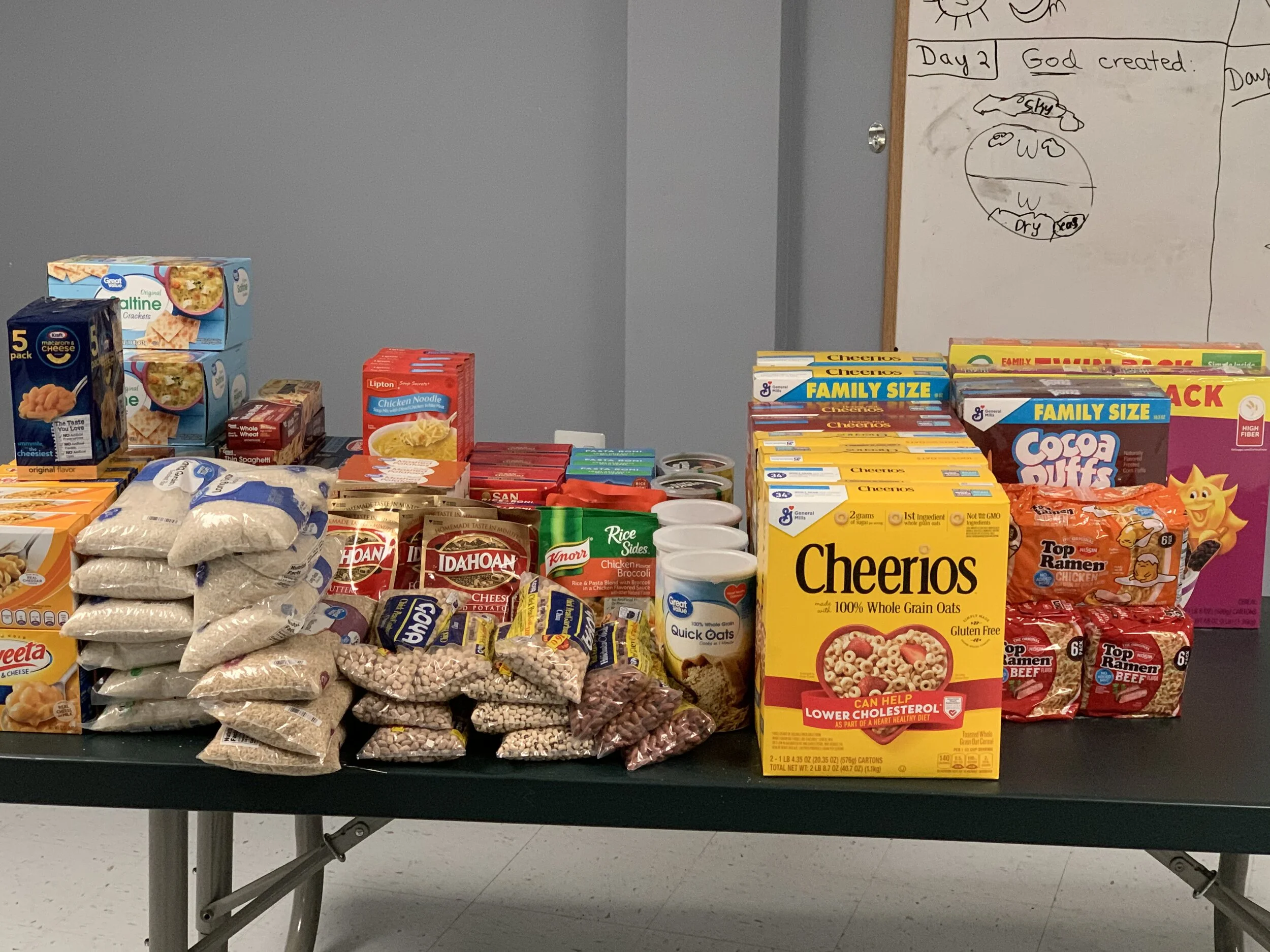 Table filled with various food and grocery items including cereal boxes, rice, ramen, macaroni, canned goods, pasta, and snack packs, set against a wall with handwritten notes and drawings.