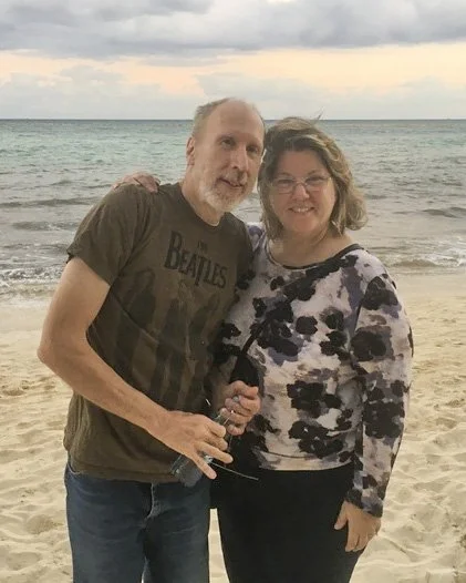 Photographer Bill Morley and his lovely wife, Karen standing on the beach near the ocean, smiling at the camera during sunset.