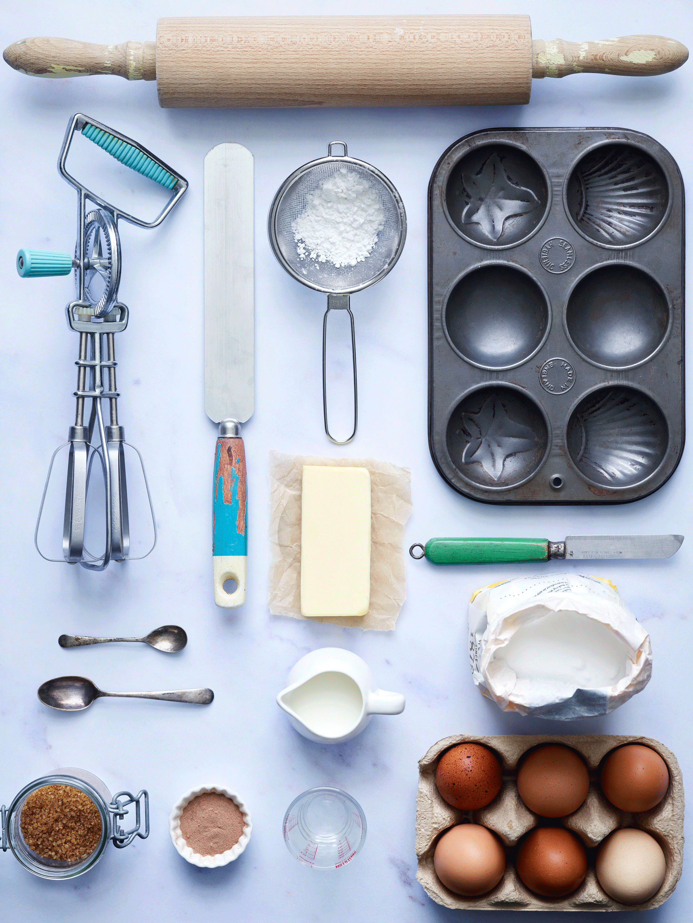 Baking ingredients and tools arranged on a table. Includes a rolling pin, hand mixer, offset spatula, flour sifter with powdered sugar, butter, muffin tin, two spoons, milk in a jug, brown sugar, cocoa powder, measuring cup, six eggs in a carton, flo