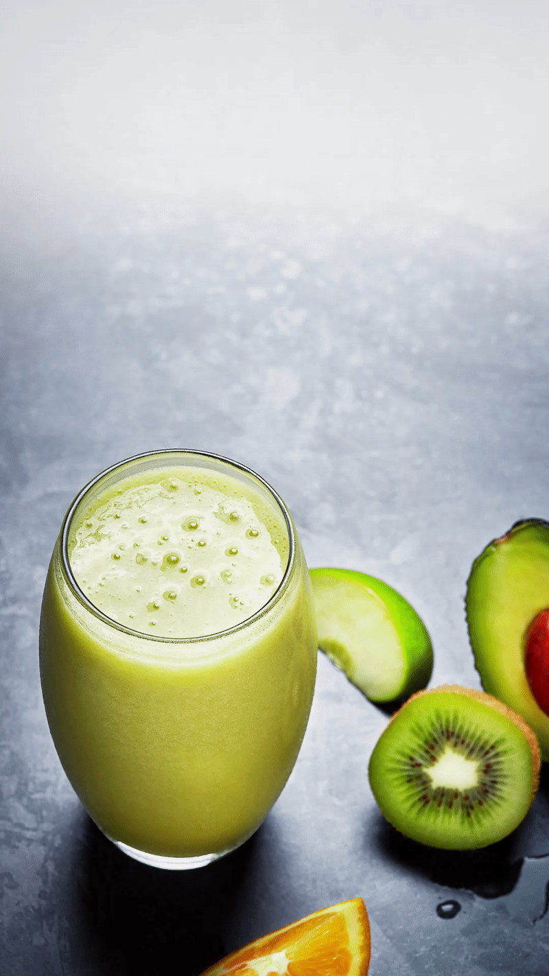 Green smoothie in glass with slices of kiwi, avocado, and orange on dark surface.