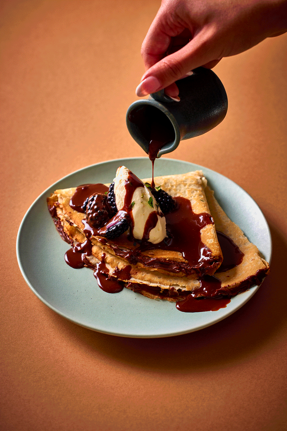 Crepes topped with ice cream, blackberries, and chocolate sauce being poured from a jug on a plate.