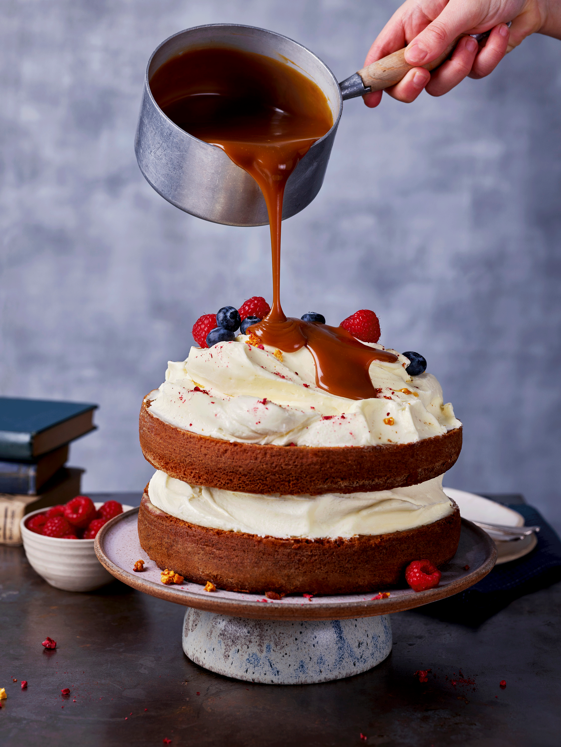 Layered cake with white frosting and berries, being drizzled with caramel sauce from a saucepan.