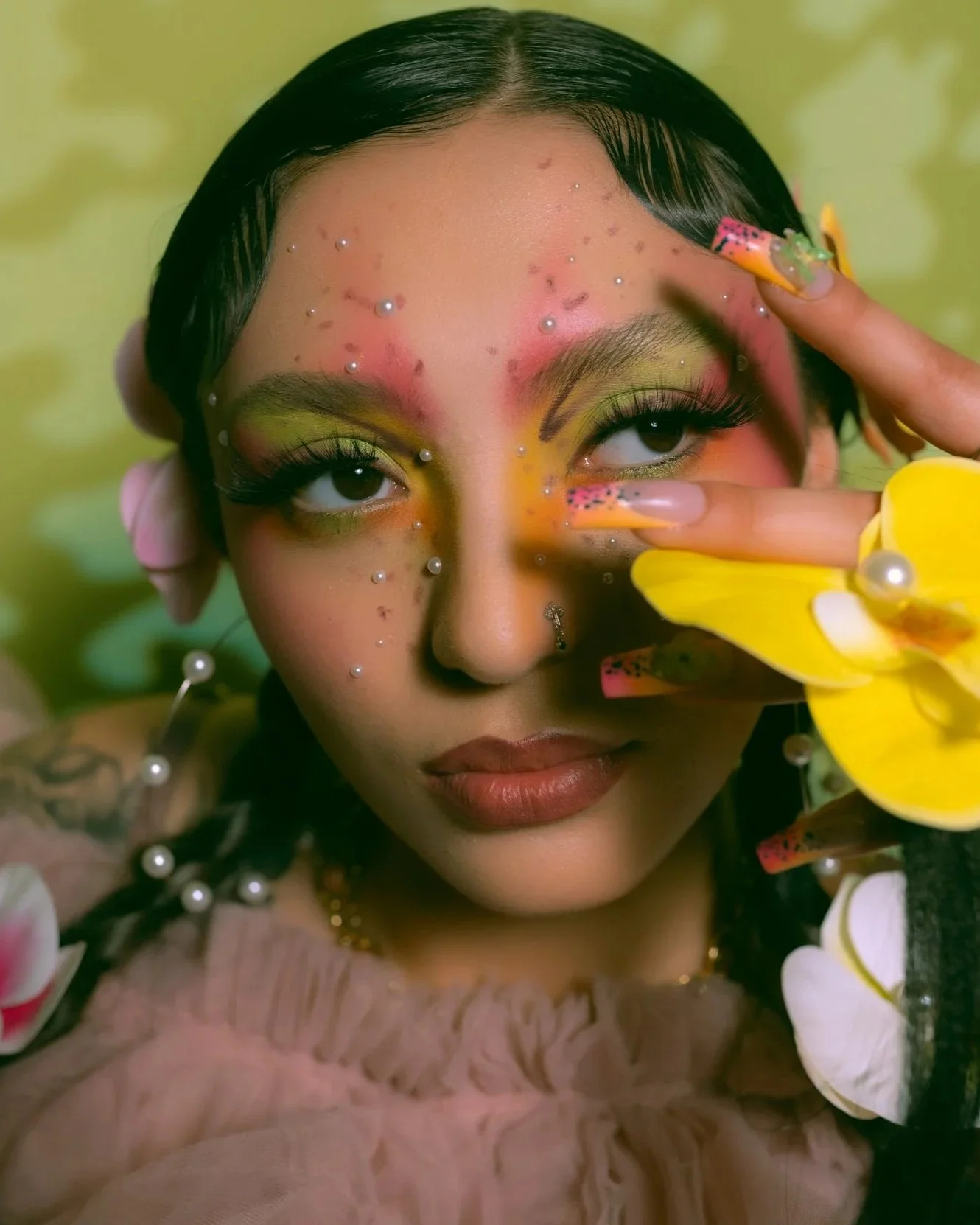 A close-up portrait of a young woman with elaborate makeup and accessories, holding yellow and white flowers near her face, with a green background.