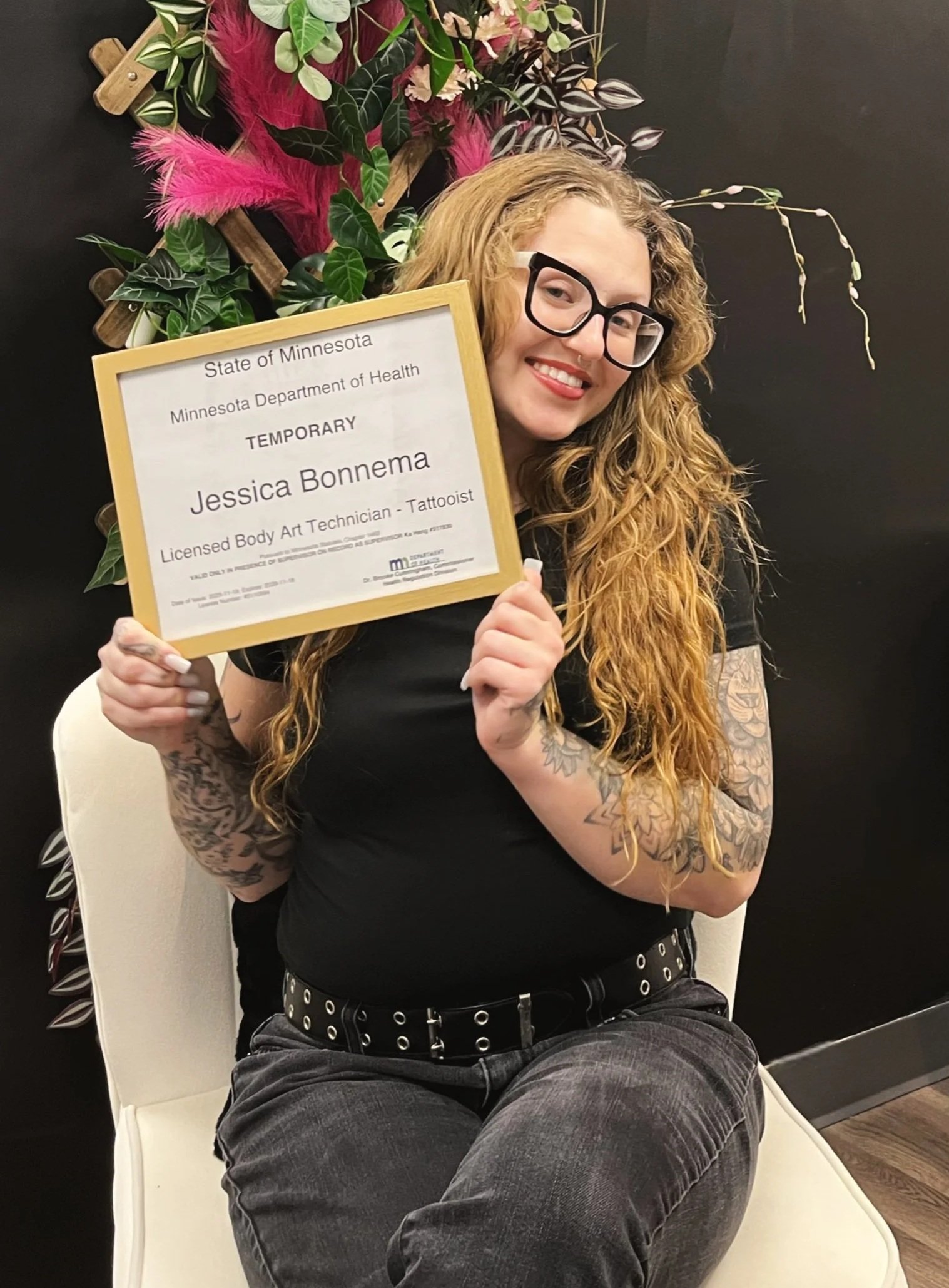 A woman with long red hair, glasses, and tattoos is smiling while sitting on a white chair, holding a certificate for Jessica Bonnema, a licensed body art technician and tattooist. She is wearing a black shirt and gray pants, and there is a black wall with pink and green decorative plants in the background.