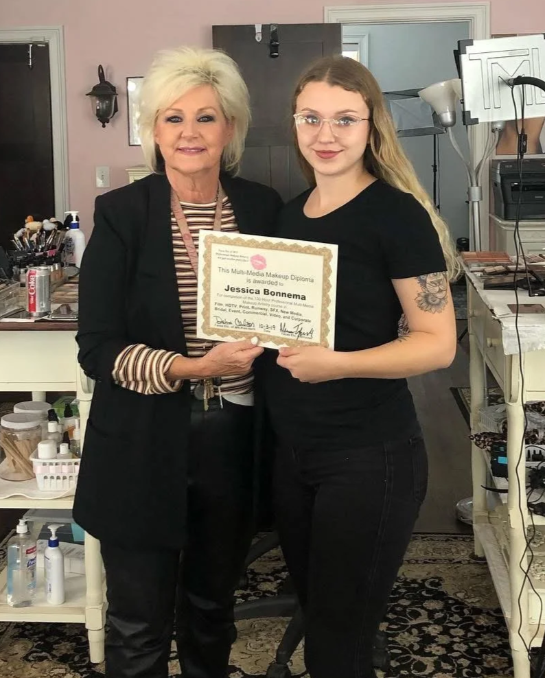 Two women standing together in a makeup studio, holding a diploma. The woman on the left has short blonde hair, is wearing a black blazer and striped shirt, while the woman on the right has long blonde hair, glasses, and a tattoo on her arm, wearing a black shirt.