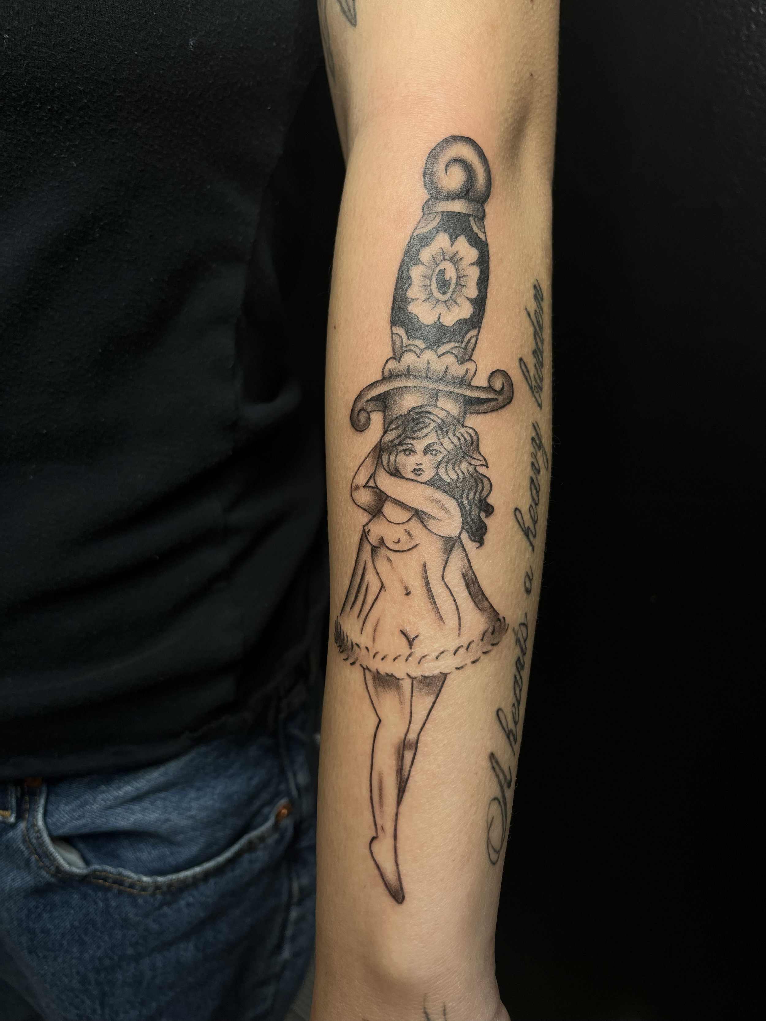 Tattoo of a woman with long hair in a short dress, obscuring one eye, with a large dagger or sword positioned vertically above her head. There is cursive text next to the tattoo.