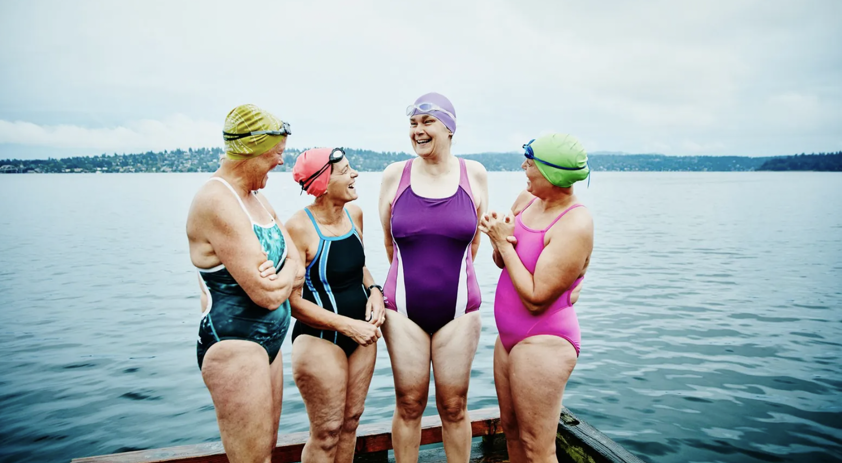 Group of women of various ages in swimsuits laughing and standing in front of a lake