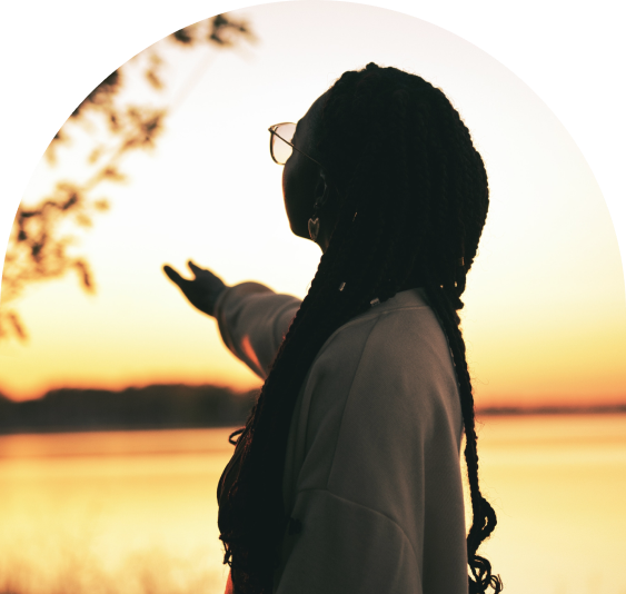 A woman with dreadlocks wearing glasses and a light-colored jacket gestures towards a sunset over a body of water.