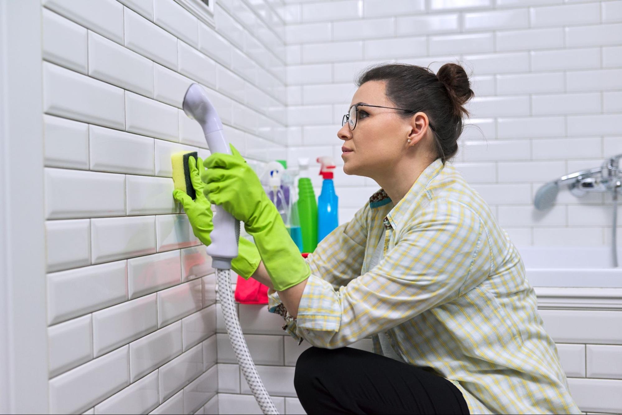A woman wearing a light yellow-checkered long-sleeved shirt, black leggings, neon green latex gloves, and eyeglasses washes a white tile wall with steam. Various colored cleaners and a stainless bathtub faucet are visible.