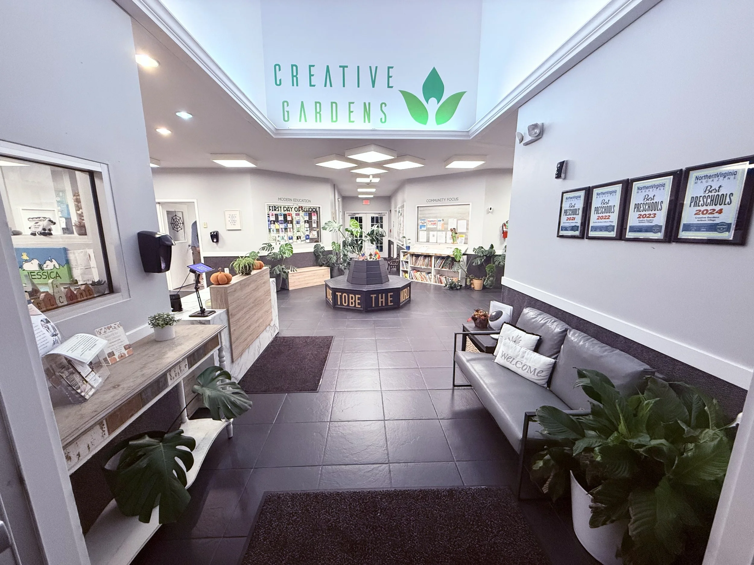 Interior of a bright community center or preschool lobby with plants, framed awards, a sitting area with pillows, a reception desk, and a sign overhead that reads 'Creative Gardens' with a leaf logo.