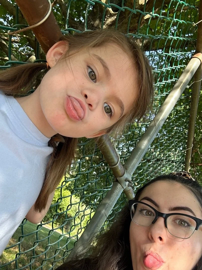 Two girls making silly faces and sticking out their tongues, standing beside a green chain-link fence outdoors.