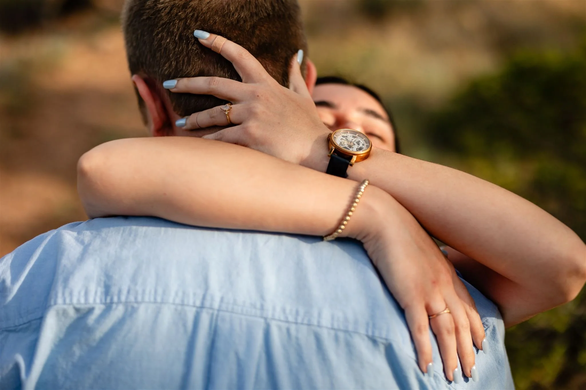 womans arms wearing watch and engagement rind hugging fiance with a blue shirt