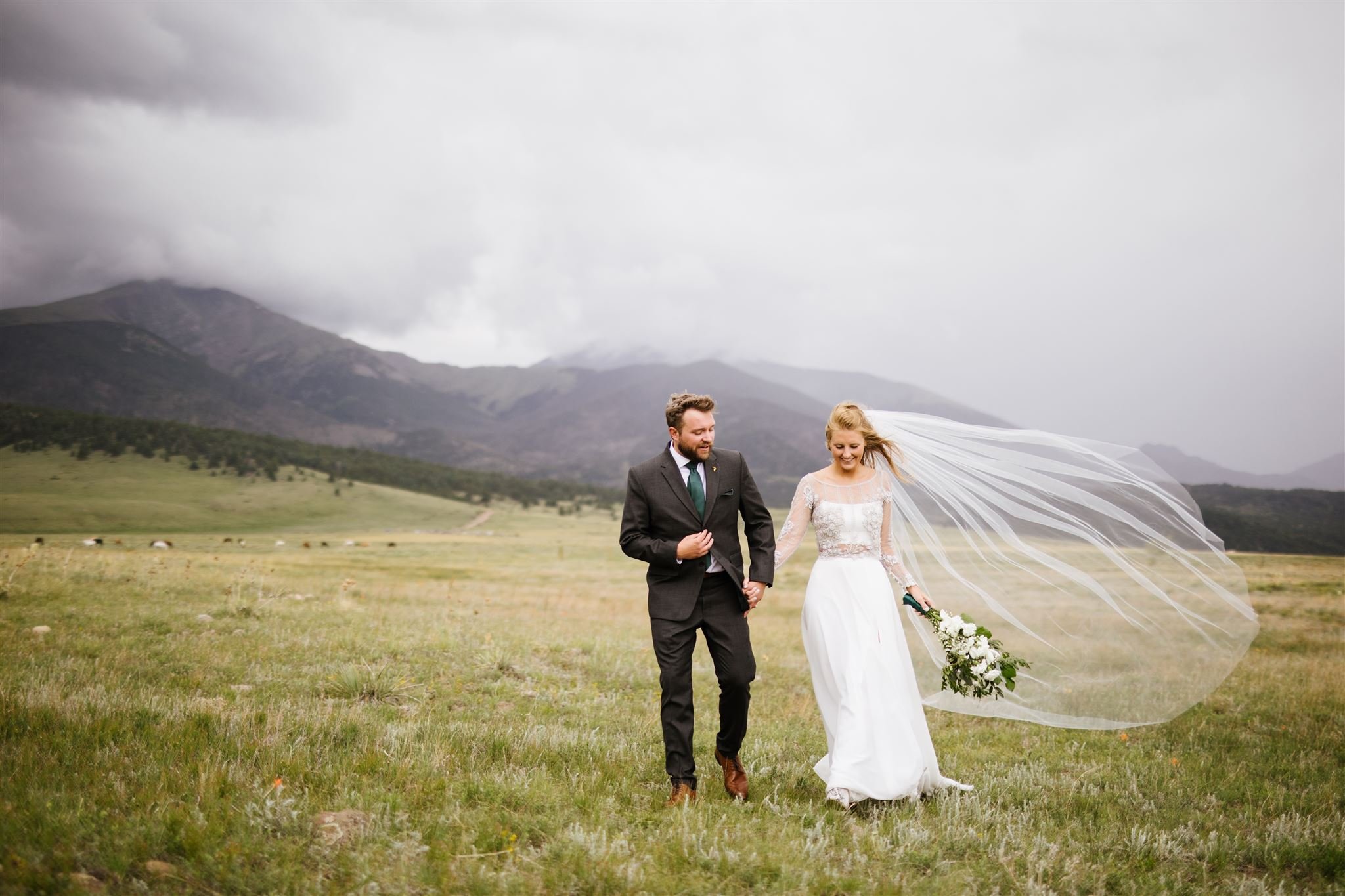 Bride and Groom walking in pasture with mountains in the background and the brides veil blowing in the wind.