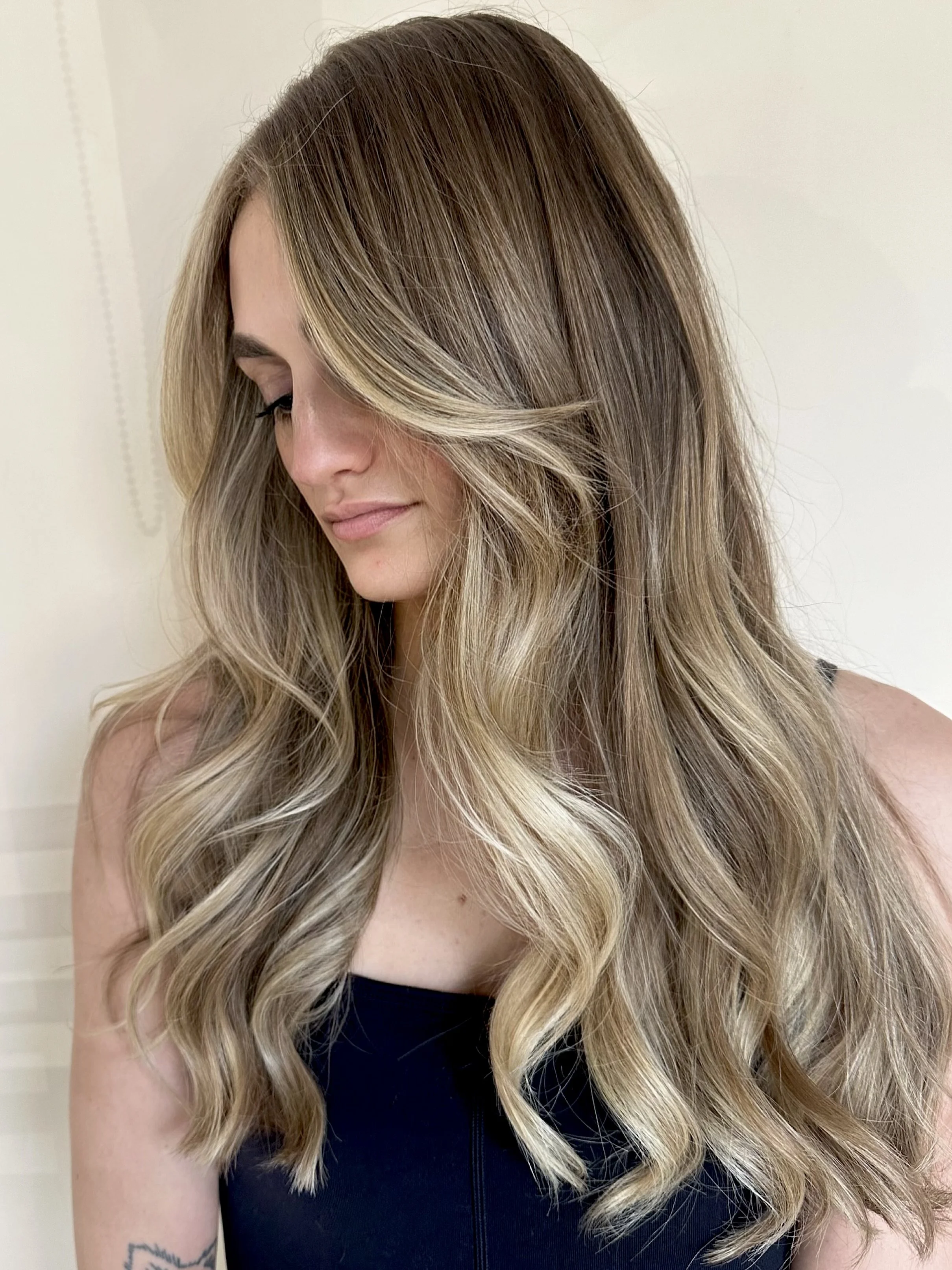 Close-up of a woman with long, wavy, blonde hair with dark roots, wearing a black top, standing against a plain light-colored wall.