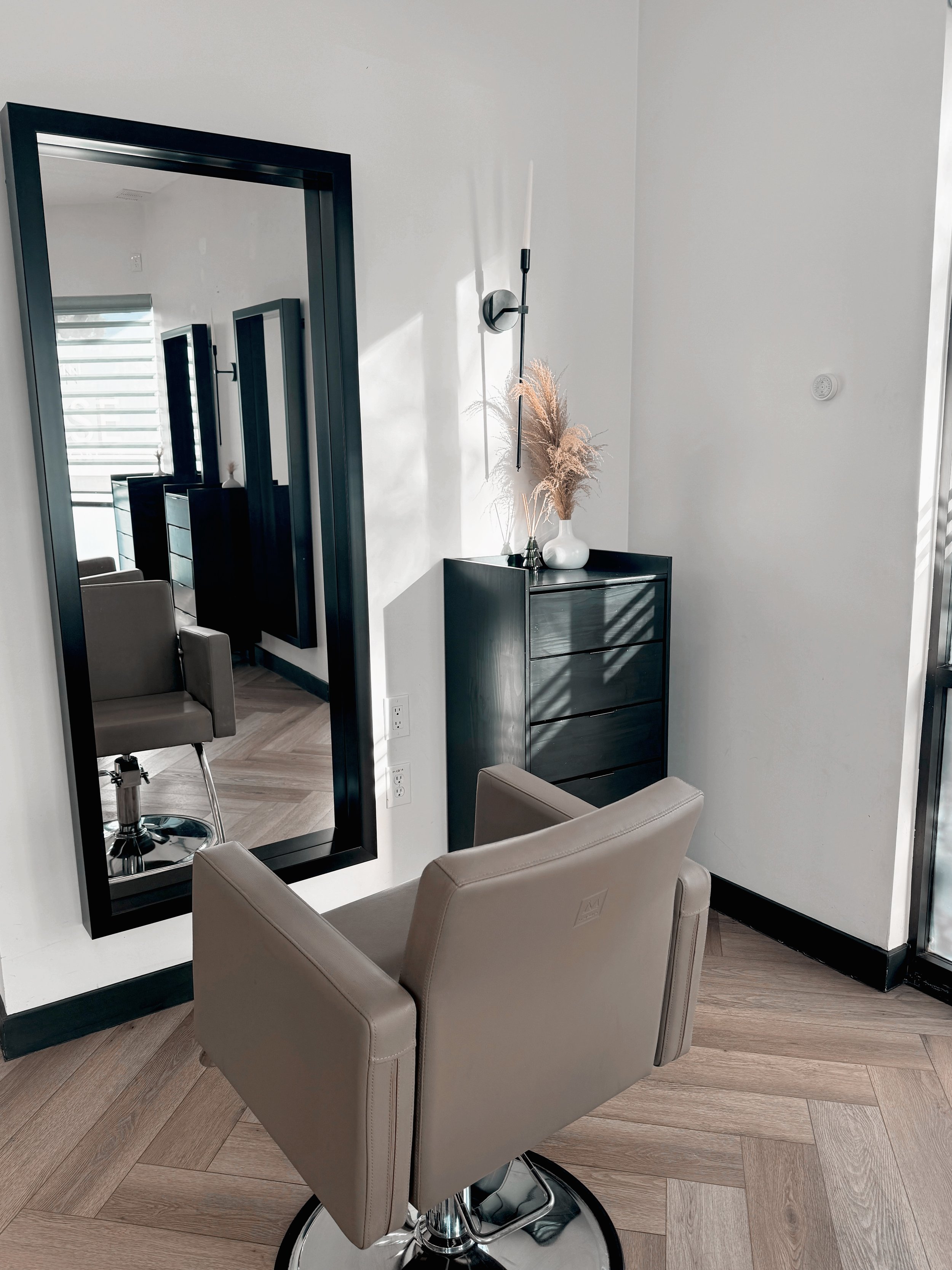 A salon chair facing a large black-framed mirror on a white wall, with a beige armchair in the foreground and a black cabinet with a white vase and dried pampas grass on top. There is a window with horizontal blinds reflected in the mirror.