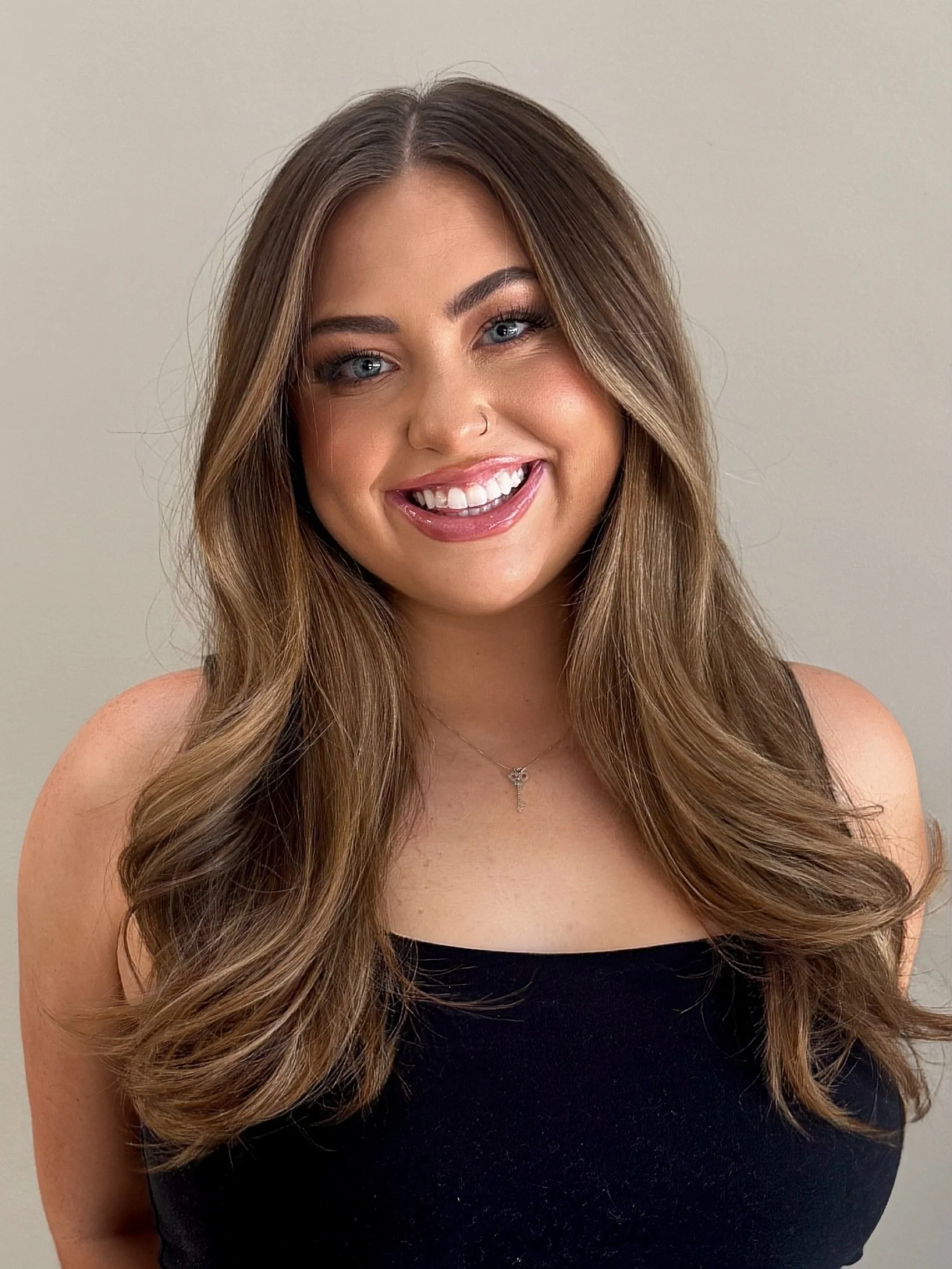 Portrait of a young woman with long wavy light brown hair, blue eyes, a nose piercing, and a bright smile. She is wearing a black top and a delicate necklace, against a plain light background.