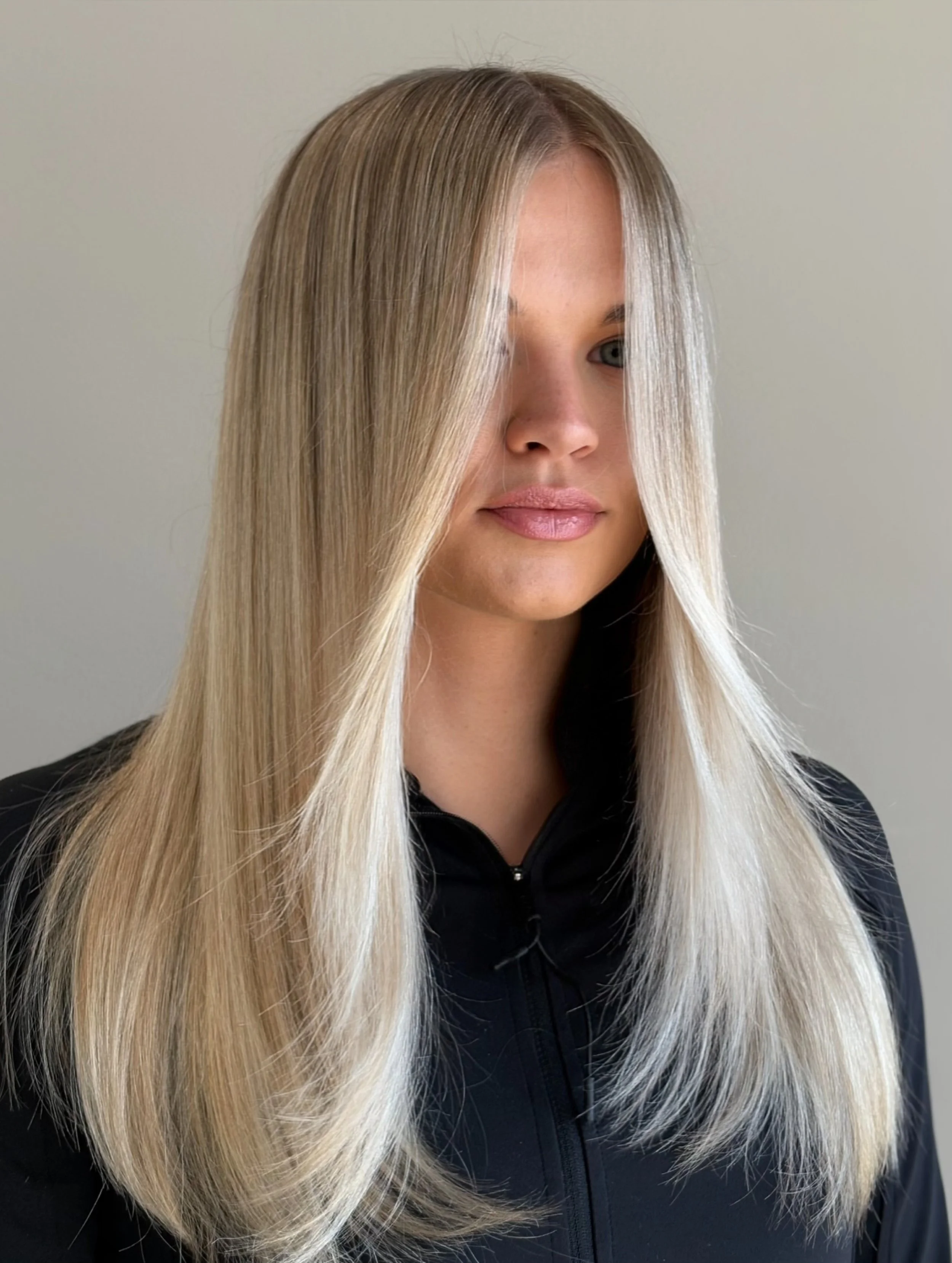 Close-up of a woman with long blonde hair, wearing a black zip-up top, standing against a plain light-colored wall.