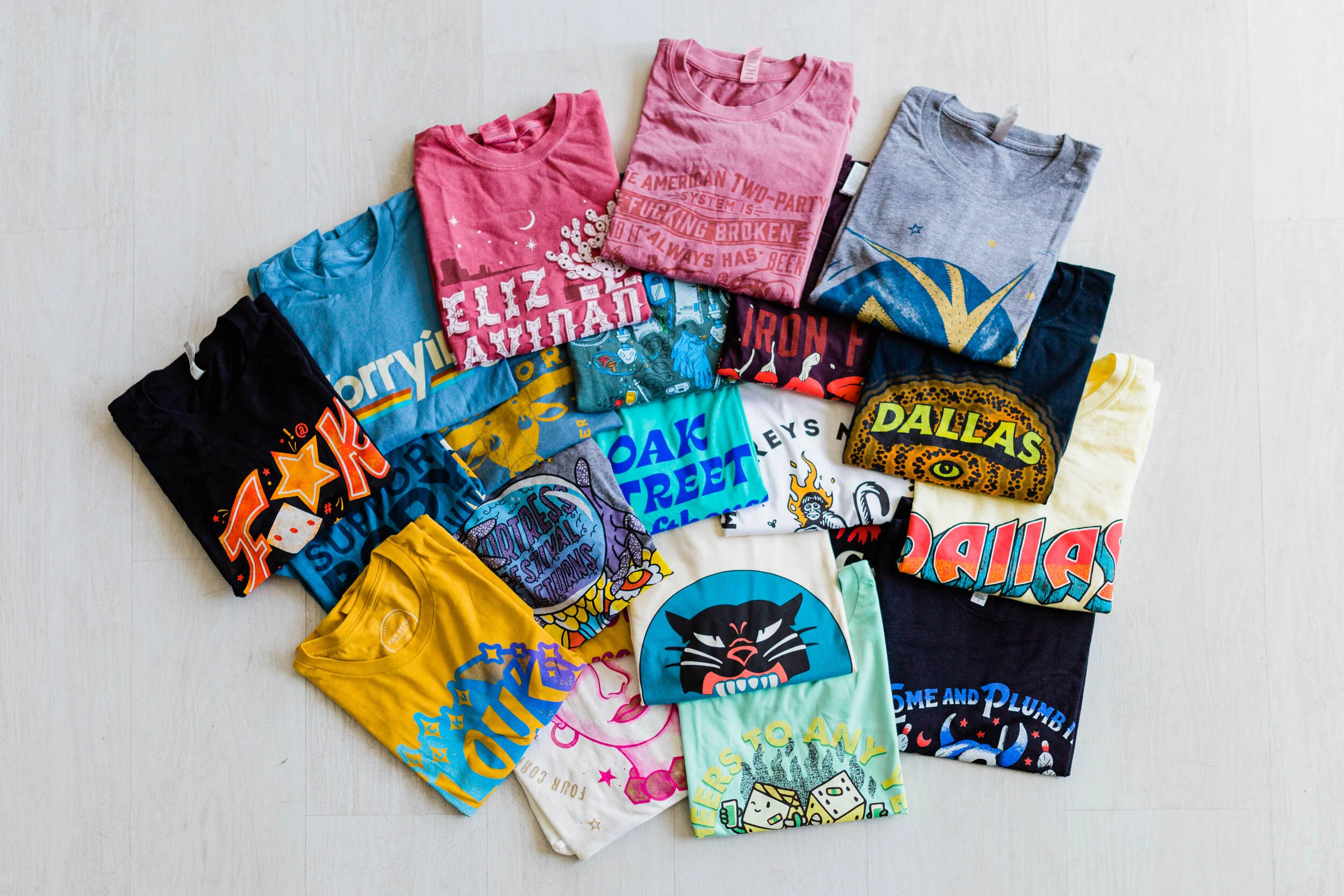 A collection of colorful, graphic T-shirts arranged on a light wooden floor, featuring various designs, logos, and characters.