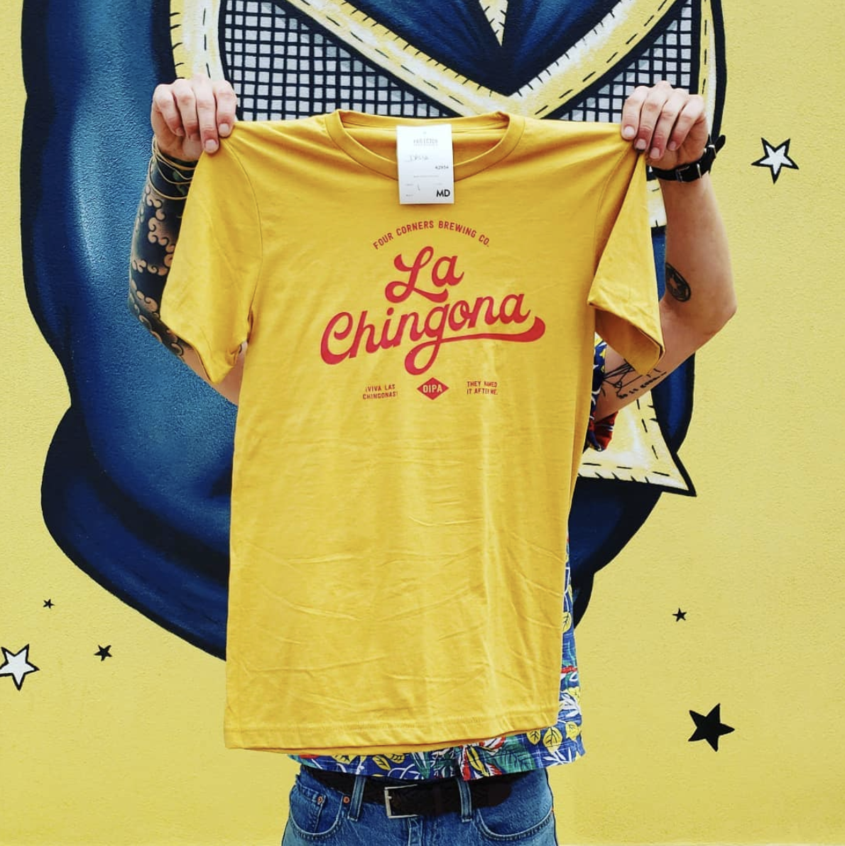 Person holding up a yellow T-shirt with red text that reads 'La Chingona' in front of a colorful mural background.