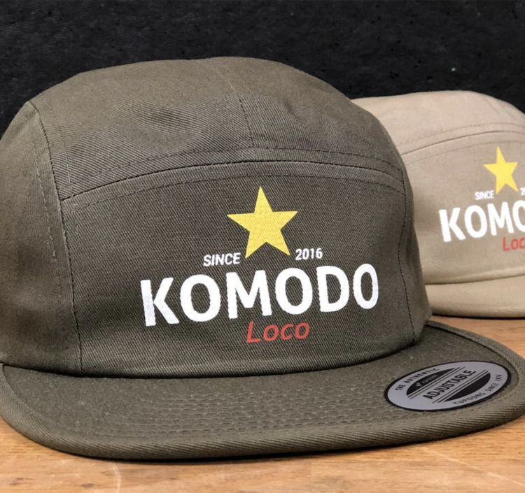 Olive green snapback cap with white and red text that reads 'Since 2016 Komodo Loco' and a yellow star above the text, placed on a wooden surface with a similar beige cap in the background.