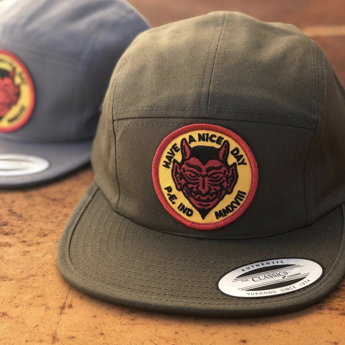 A green baseball cap with a round patch featuring a devil face, the words 'LIVE A NICE DAY PEACE AND LOVE' embroidered around the patch, and a similar blue cap in the background. The cap has a sticker that reads 'Authentic The Classics Yupoong Since 