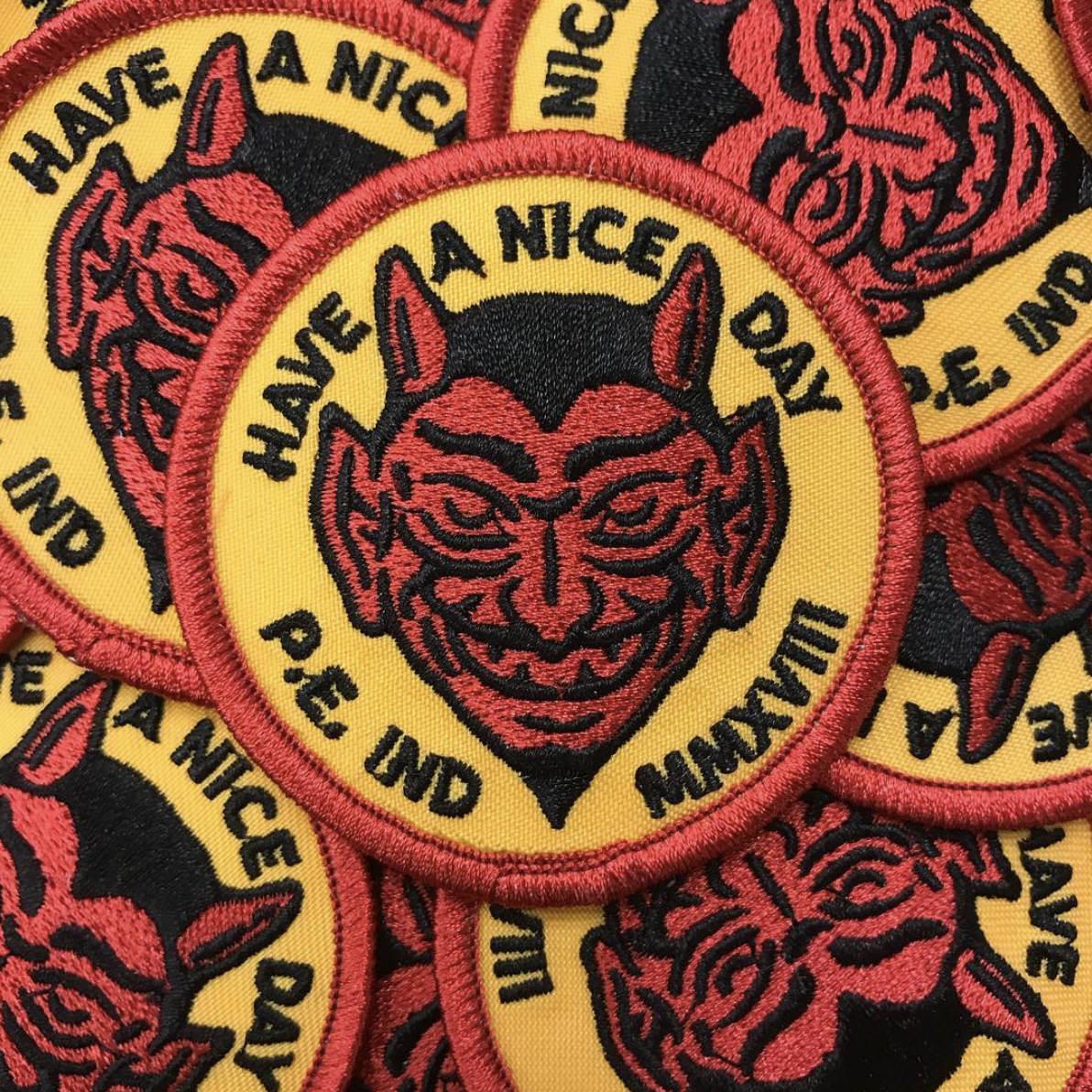 A stitched patch featuring a devil's face with horns and glasses on a yellow background, surrounded by the phrase 'Have a nice day do be mine' in black text, with a red border.