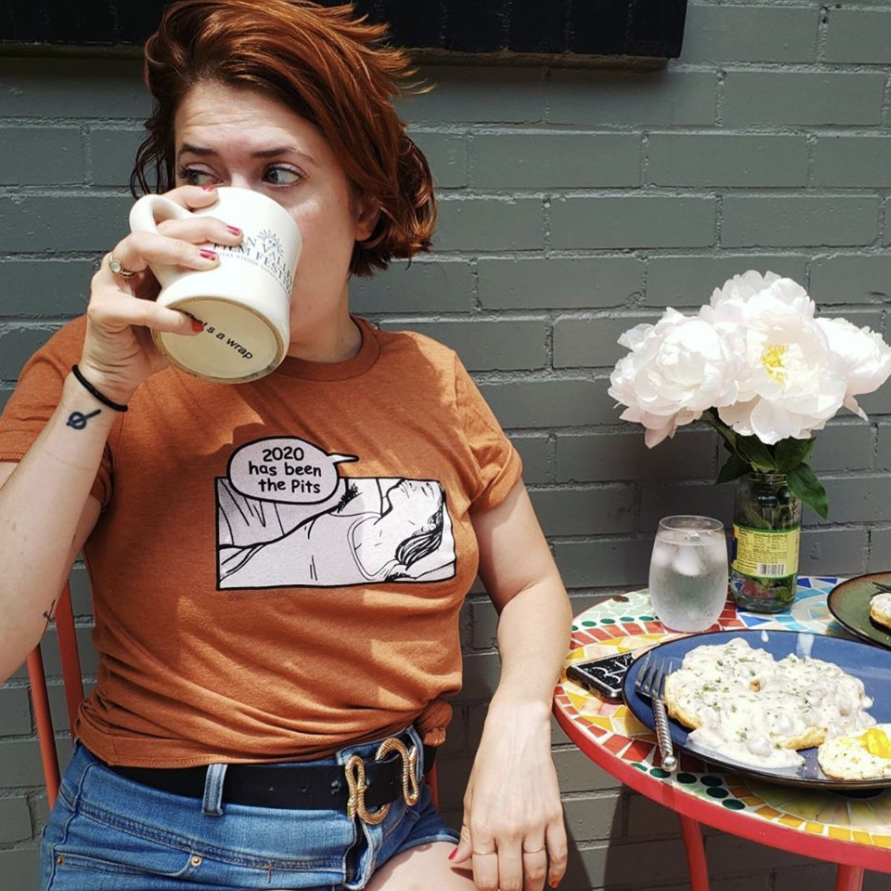 A woman with short red hair and tattoos on her left arm drinking from a white mug, sitting at a colorful table with food and a vase of white flowers against a gray brick wall.