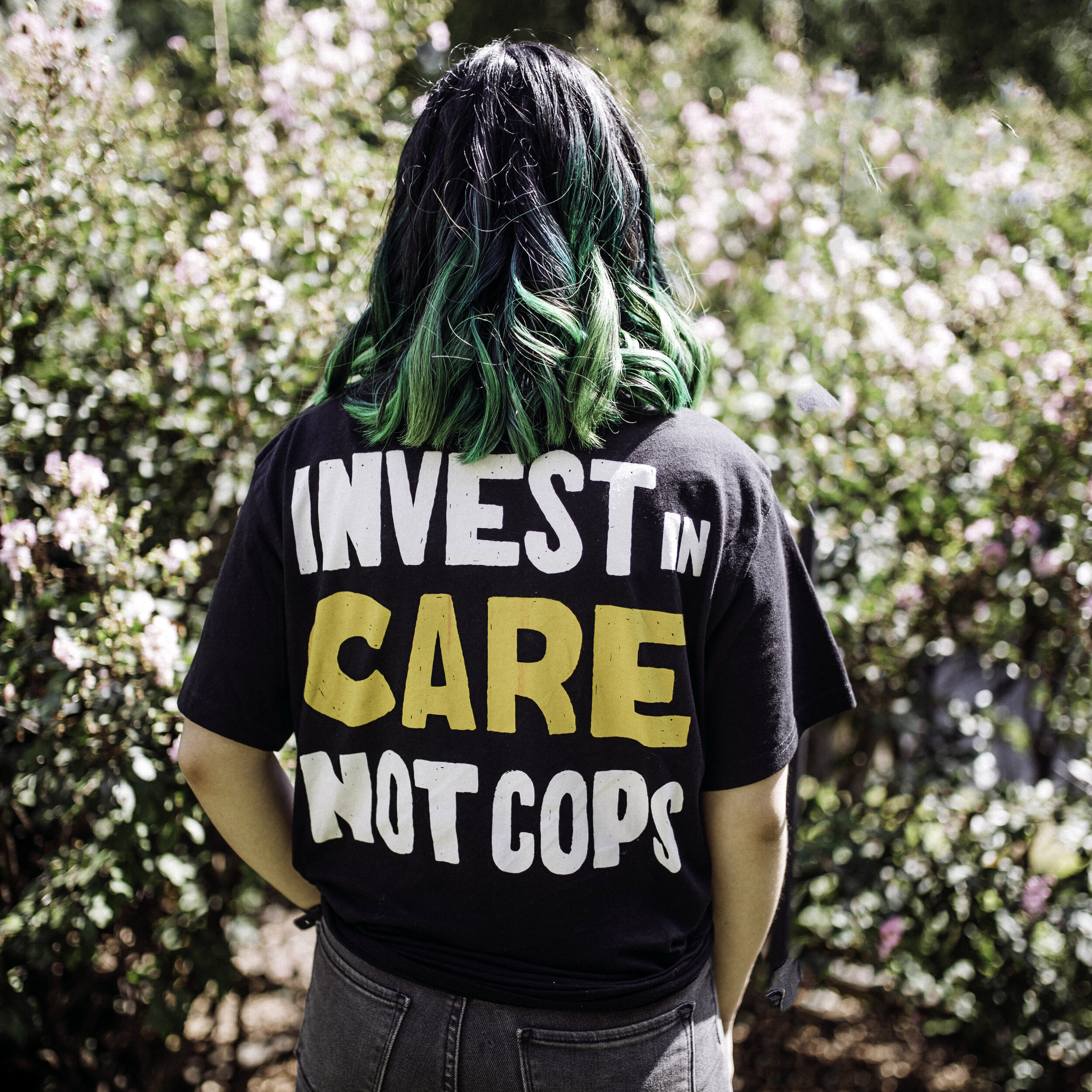 A person with shoulder-length hair dyed green and black, wearing a black t-shirt with the message 'Invest in Care, Not Cops,' standing outside in front of blooming bushes.