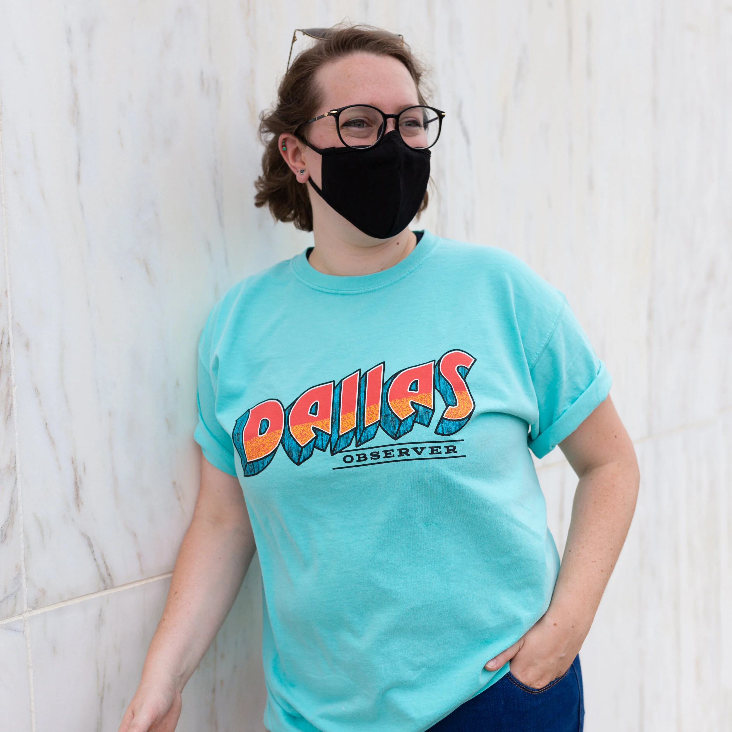 A woman with short brown hair, glasses, and a black face mask standing against a white wall. She is wearing a light blue t-shirt with the word 'Dallas' in large colorful letters and 'Observer' underneath.