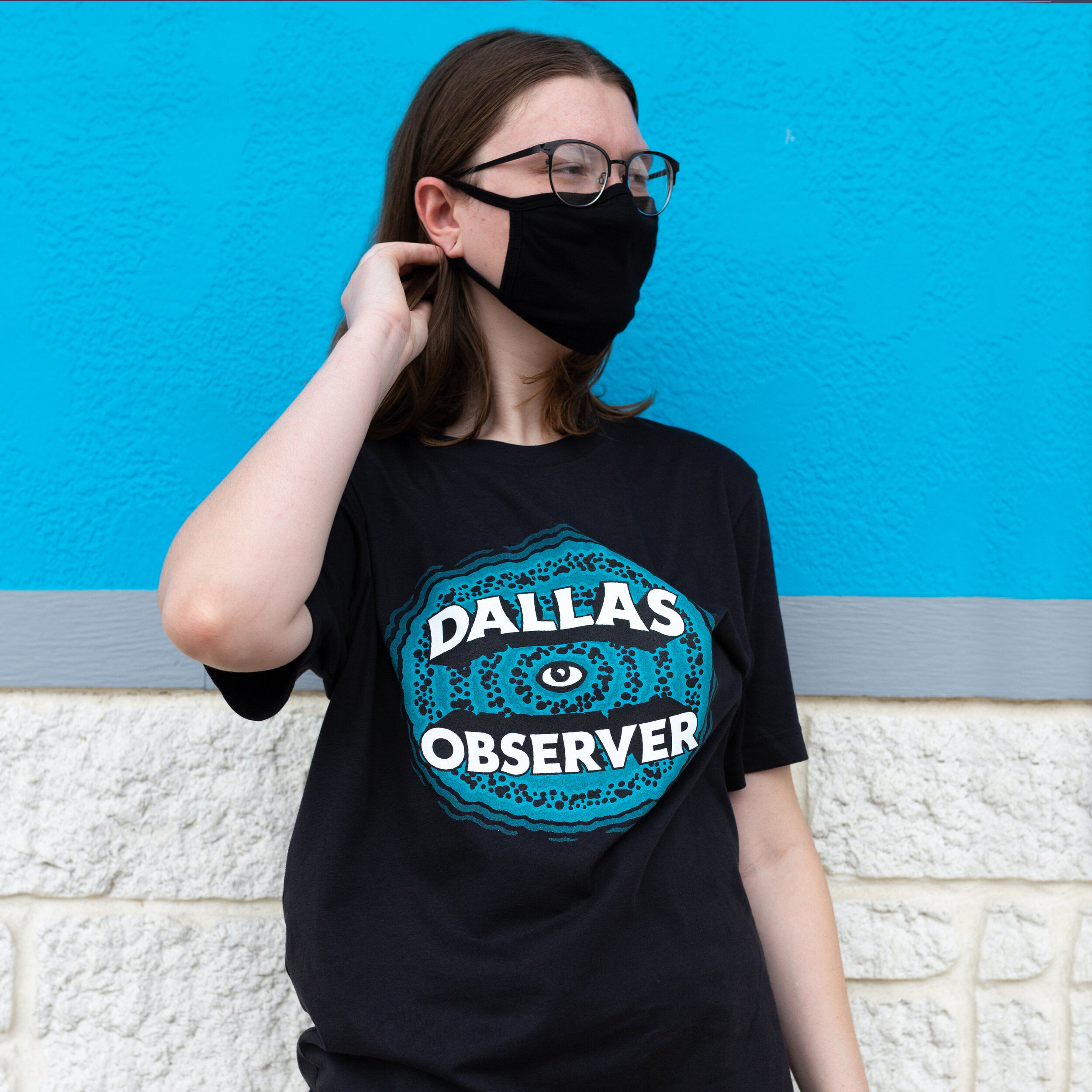 A young woman wearing glasses and a black face mask, standing against a blue wall, wearing a black T-shirt with a design that says 'Dallas Observer' with an eye graphic.