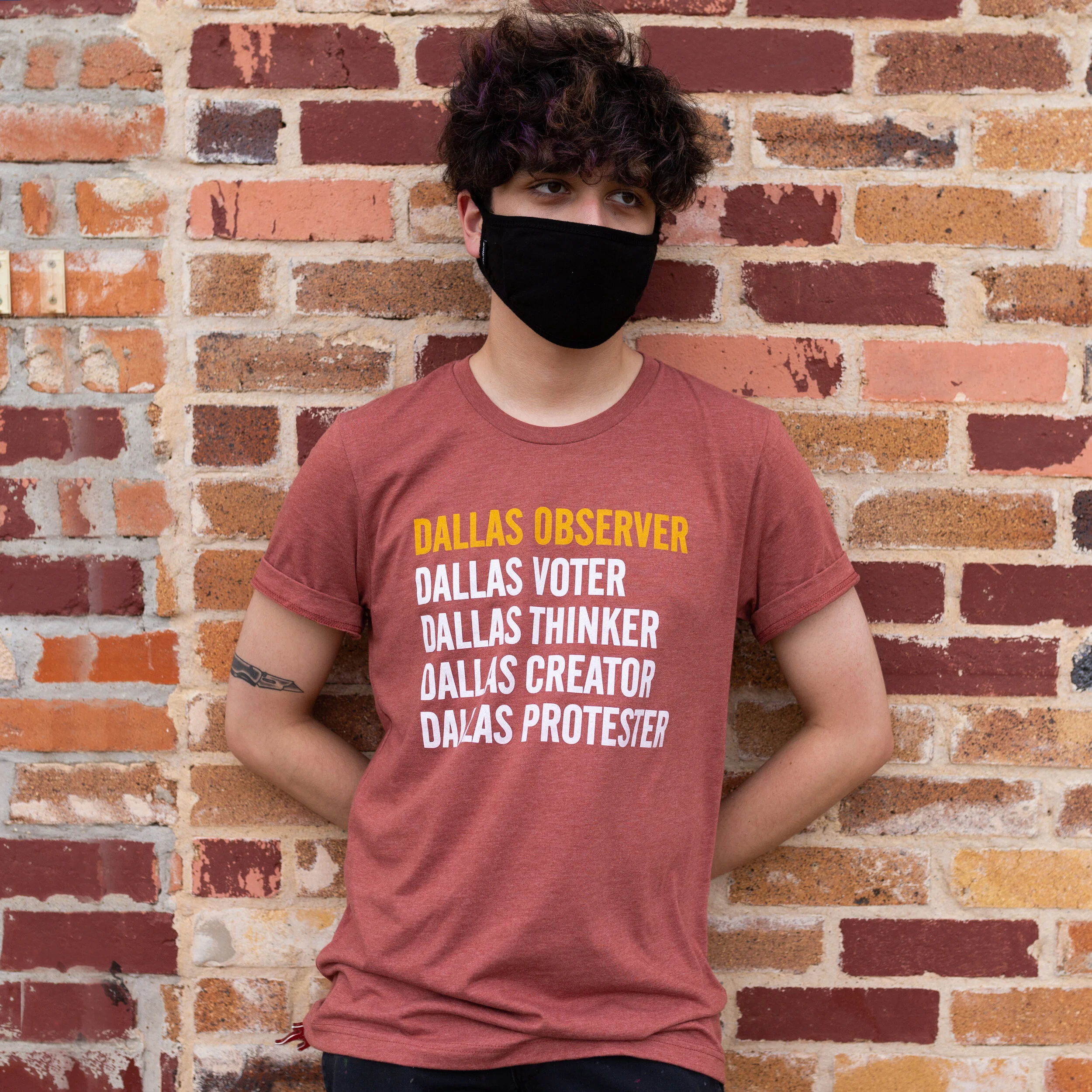 A young man with curly dark hair, wearing a black face mask and a red T-shirt with yellow and white text, standing against a brick wall.