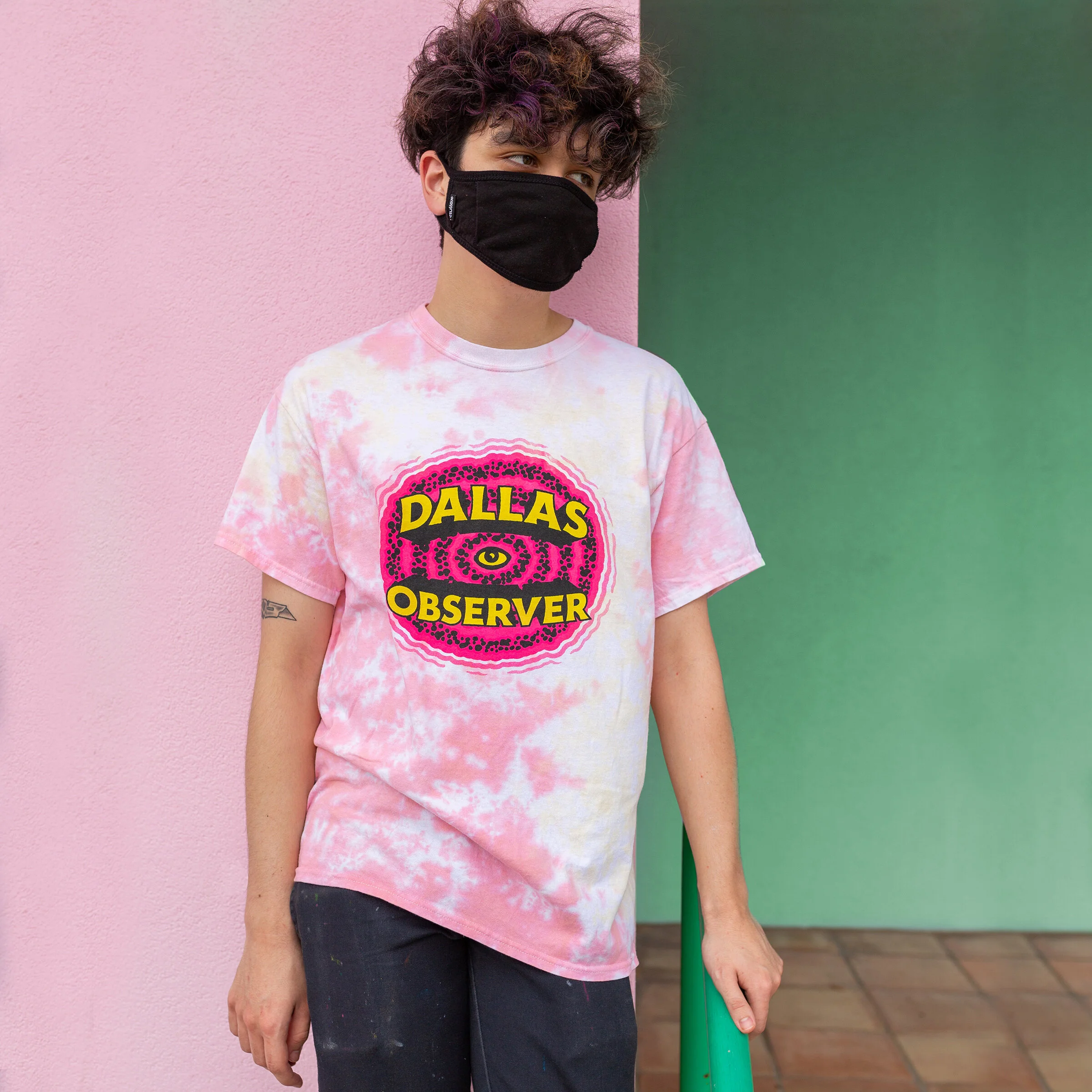 A person with curly purple hair wearing a black face mask and a pink tie-dye T-shirt with a graphic that reads 'Dallas Observer' and an eye design, standing against a background with pink and green walls.