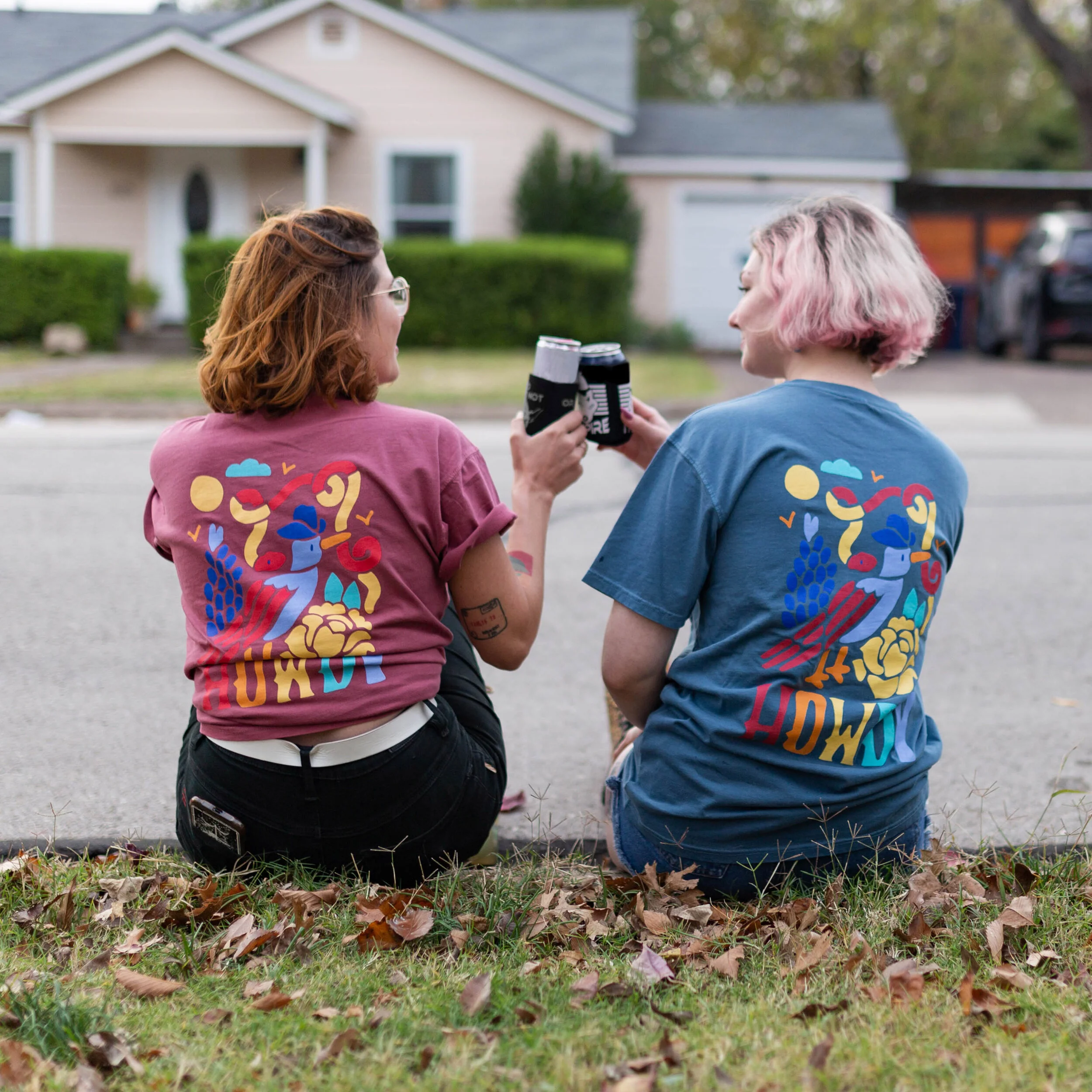 Two women with colorful T-shirts sitting on the grass on a sidewalk, holding drinks and toasting outdoors in front of a house with a hedge and trees in the background.