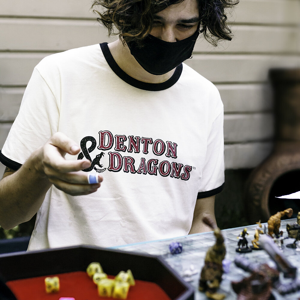 Person wearing a black face mask and a white t-shirt with purple and black text that says 'Denton & Dragons,' playing a tabletop game with miniatures and dice outdoors.