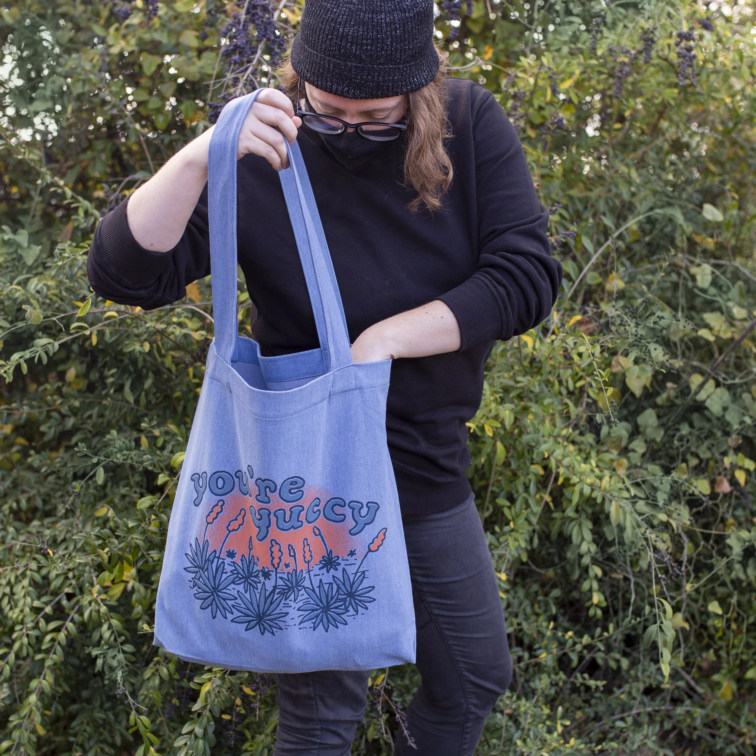 Person wearing black beanie and mask, holding a blue tote bag with the words "you're sticky" and floral illustrations, standing outdoors with green bushes in the background.