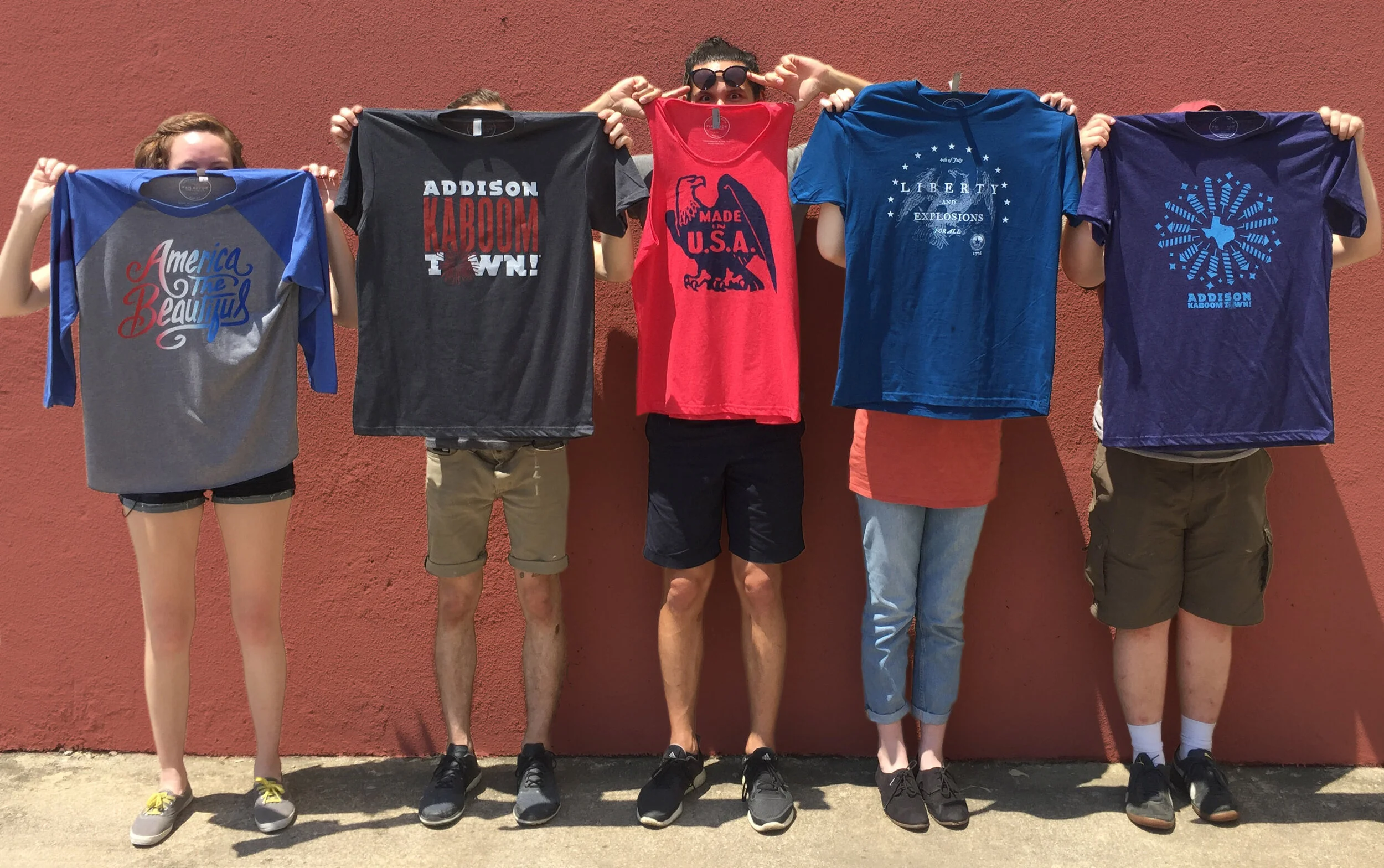 Five children standing in front of a red wall, each holding up a T-shirt in front of their face, obscuring it.