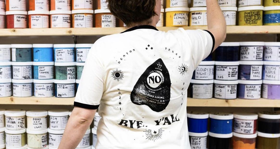 Person facing away in a store aisle with shelves of colorful paint cans, wearing a white t-shirt with black trim that has a graphic and text.
